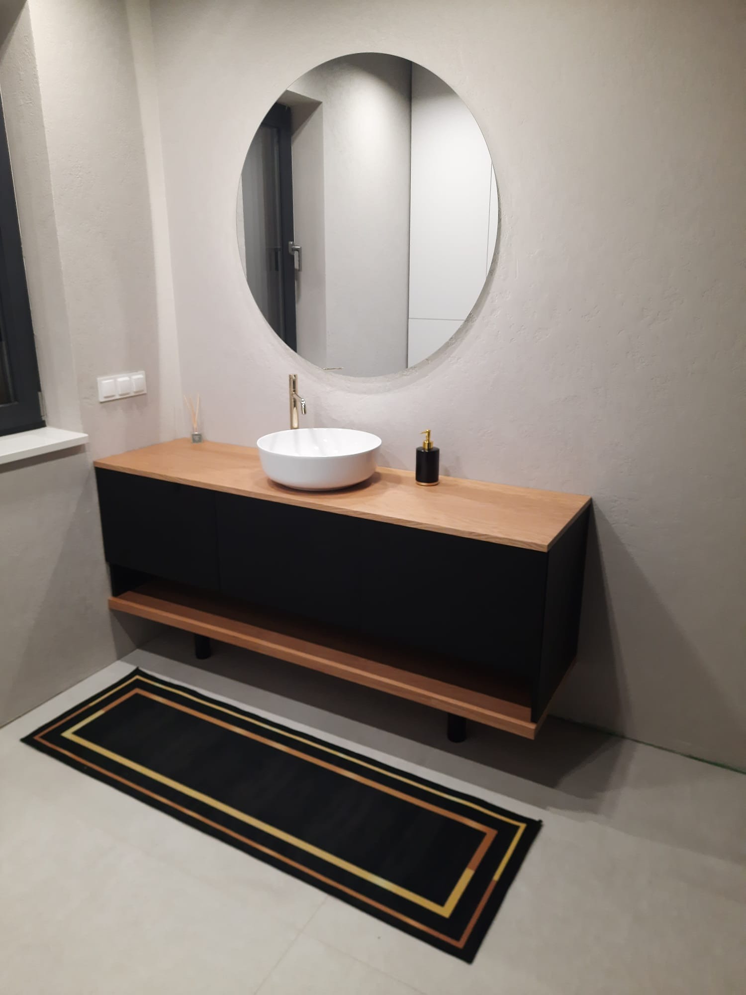 Nowoczesna łazienka z okrągłym lustrem nad szafką z umywalką nablatową. Czarna szafka z drewnianym blatem, dywanik. Minimalistyczny design, złote akcenty.