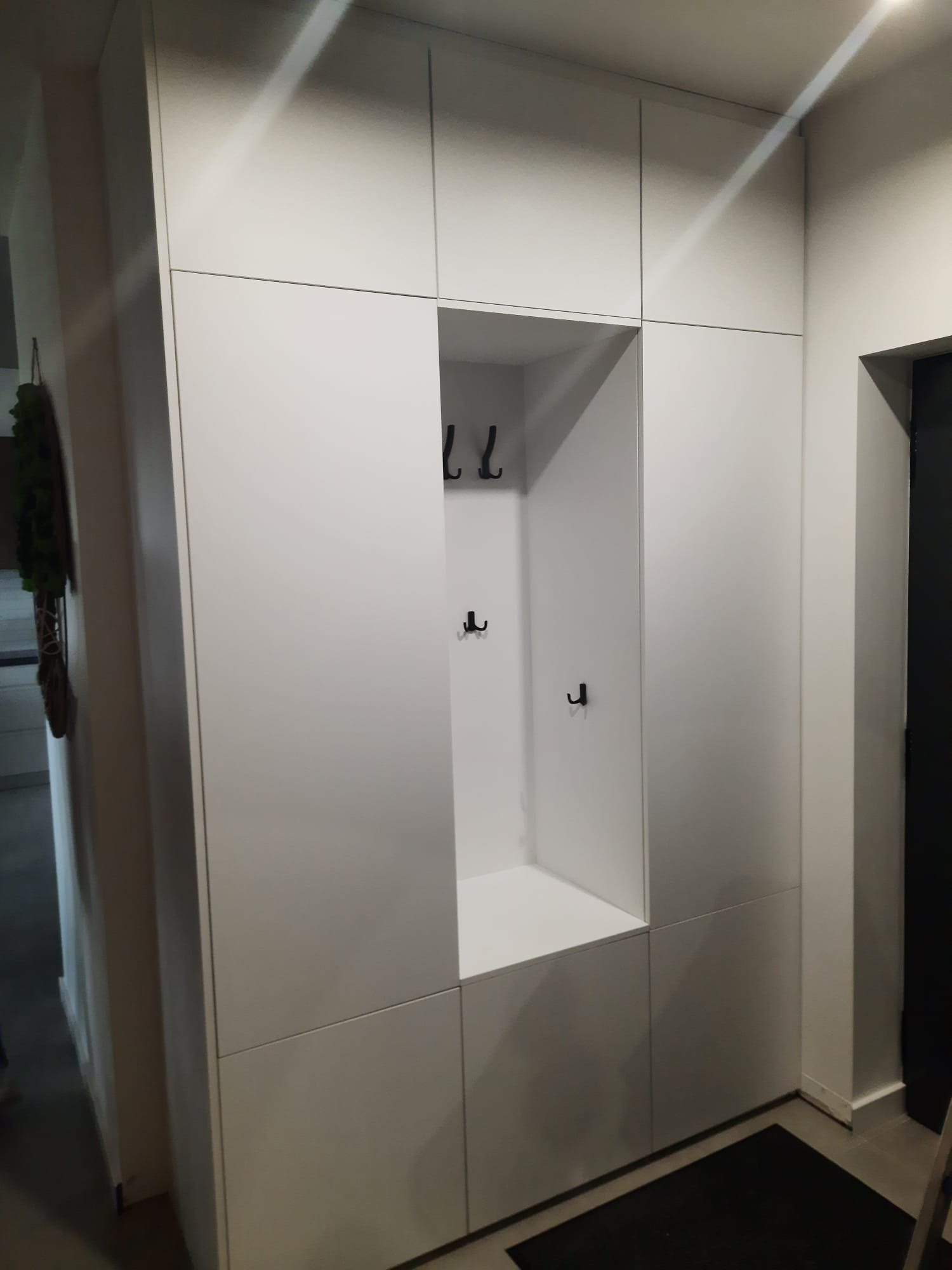 Biała szafa wnękowa z wieszakami w przedpokoju, minimalistyczny design, czarny dywanik. Widoczne oświetlenie sufitowe i fragment drzwi.