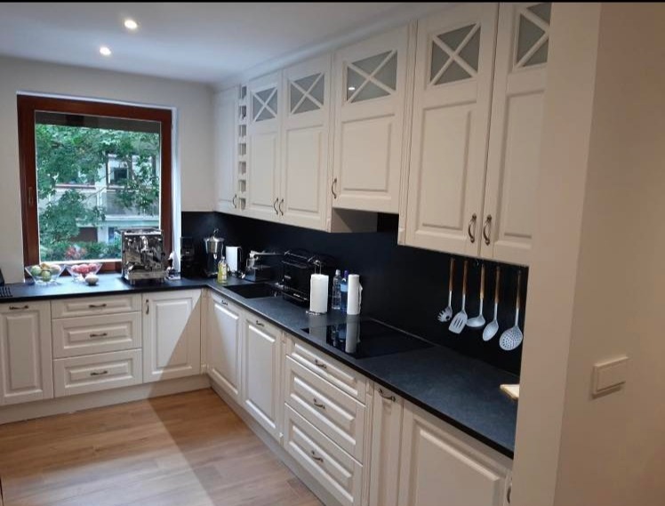 Biała, klasyczna kuchnia z frezowanymi frontami i czarnym blatem z konglomeratu kwarcowego. Nad blatem wiszące szafki z dekoracyjnymi szybkami.