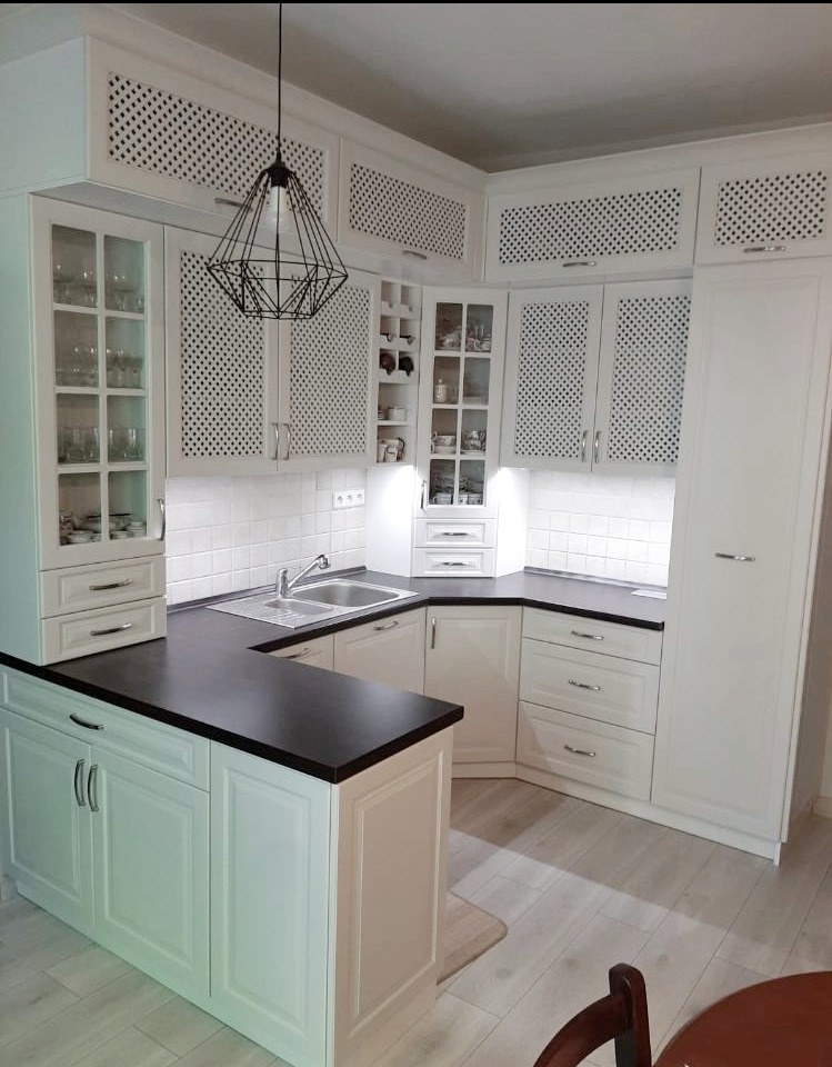 Jasna kuchnia w stylu klasycznym z czarnym blatem i ażurowymi frontami szafek. Nad wyspą kuchenną wisi nowoczesna, geometryczna lampa.