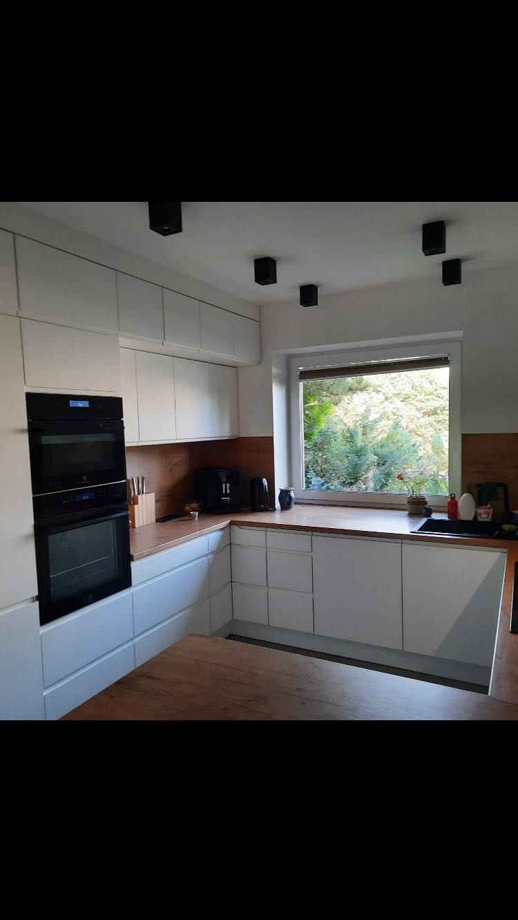 Nowoczesna kuchnia z białymi frontami, drewnianym blatem i czarnymi lampami sufitowymi. Widok na zieleń za oknem.