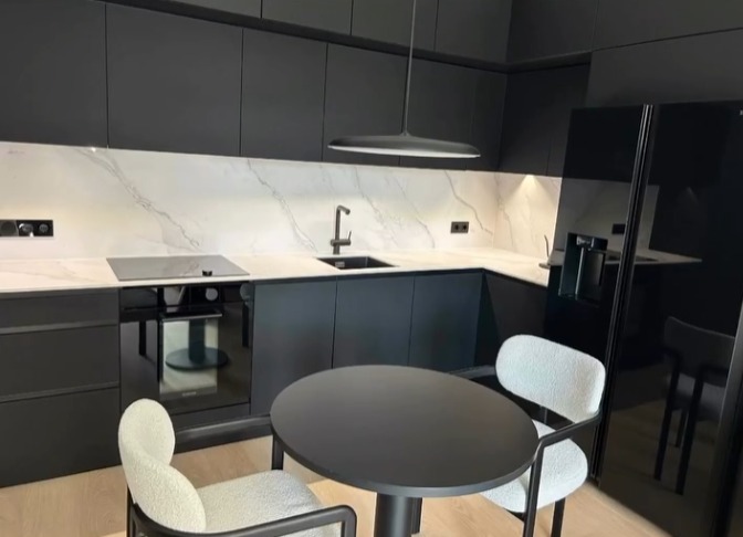 Nowoczesna, czarna kuchnia z białym blatem imitującym marmur, okrągły stół i krzesła w minimalistycznym stylu. Całość oświetlona przez wiszącą lampę.