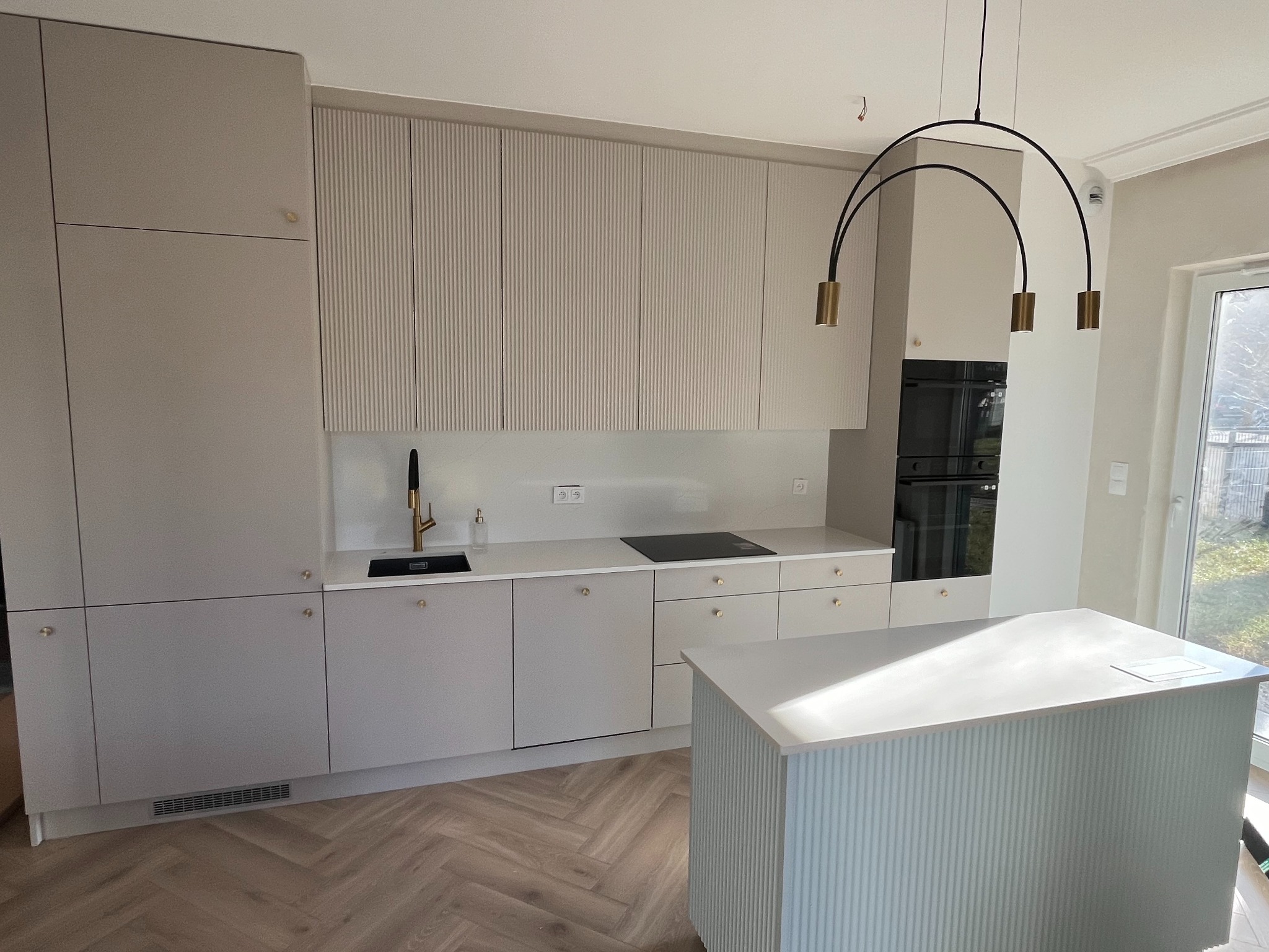 Beżowa kuchnia z ryflowanymi frontami i wyspą, złota armatura, parkiet w jodełkę. Nowoczesne oświetlenie nad wyspą.