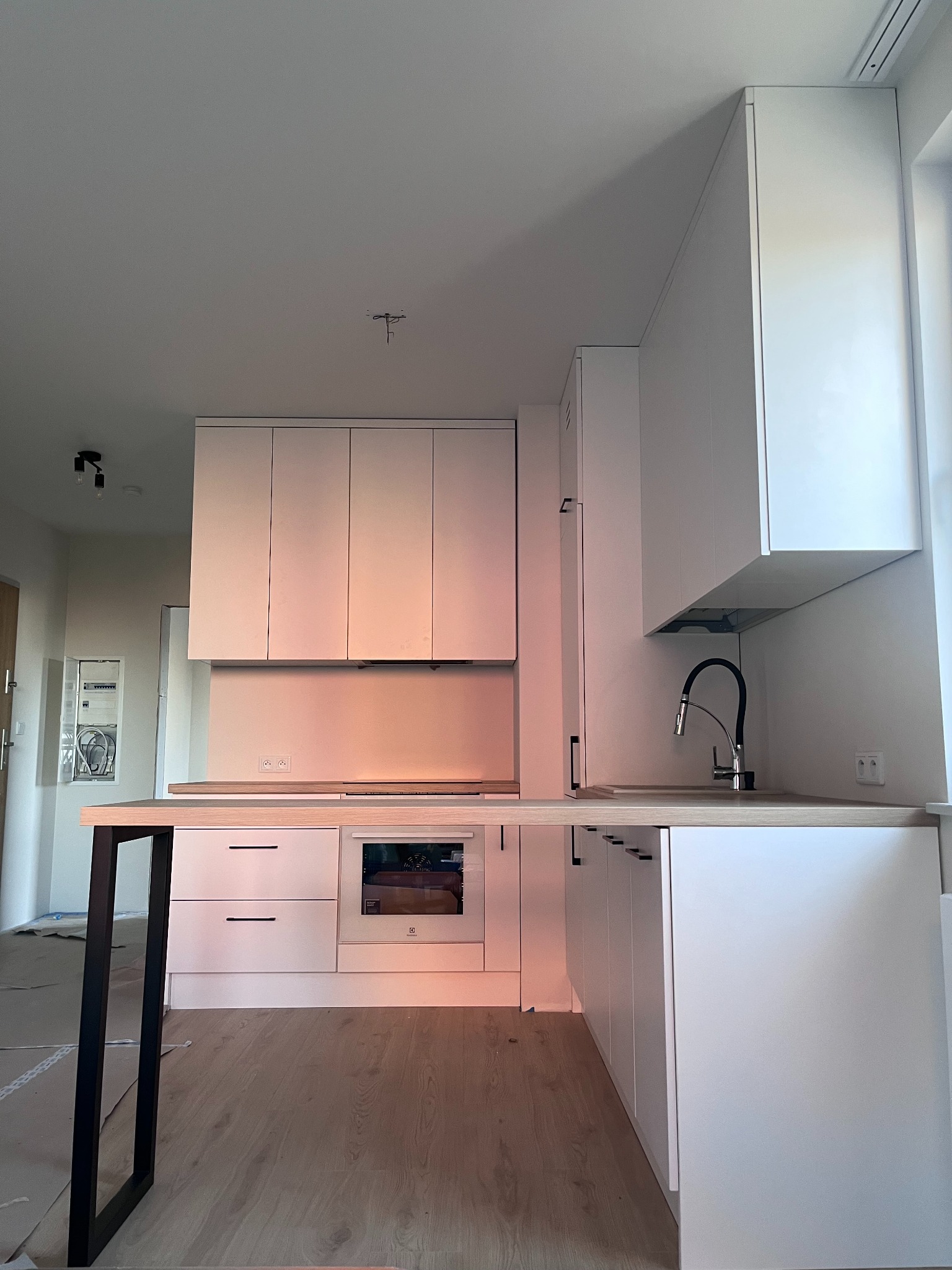 Nowoczesna, biała kuchnia z drewnianym blatem i czarną armaturą. Minimalistyczne fronty szafek, oświetlenie LED. Widoczny fragment podłogi wyłożonej panelami.