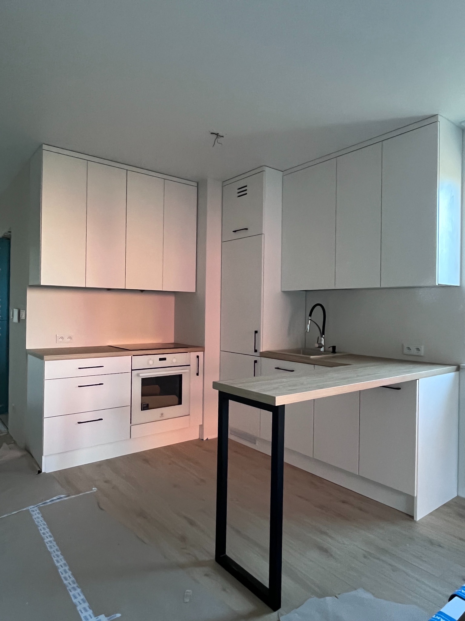 Jasna kuchnia z białymi szafkami i blatem w odcieniach drewna. Minimalistyczny design, czarne uchwyty. Na podłodze zabezpieczone panele. W tle widoczne gniazdka elektryczne.