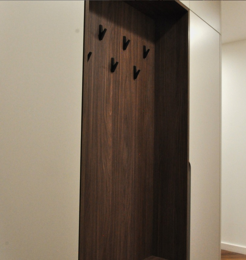 Wnękowa szafa z ciemnego drewna z wieszakami na ubrania, wkomponowana w jasną zabudowę. Minimalistyczny design, widoczne detale wykończenia.