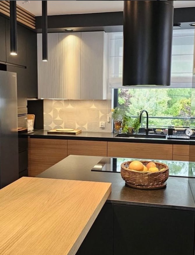 Nowoczesna kuchnia z wyspą, czarnymi i drewnianymi frontami, oświetleniem punktowym, wyciągiem i widokiem na zieleń za oknem.