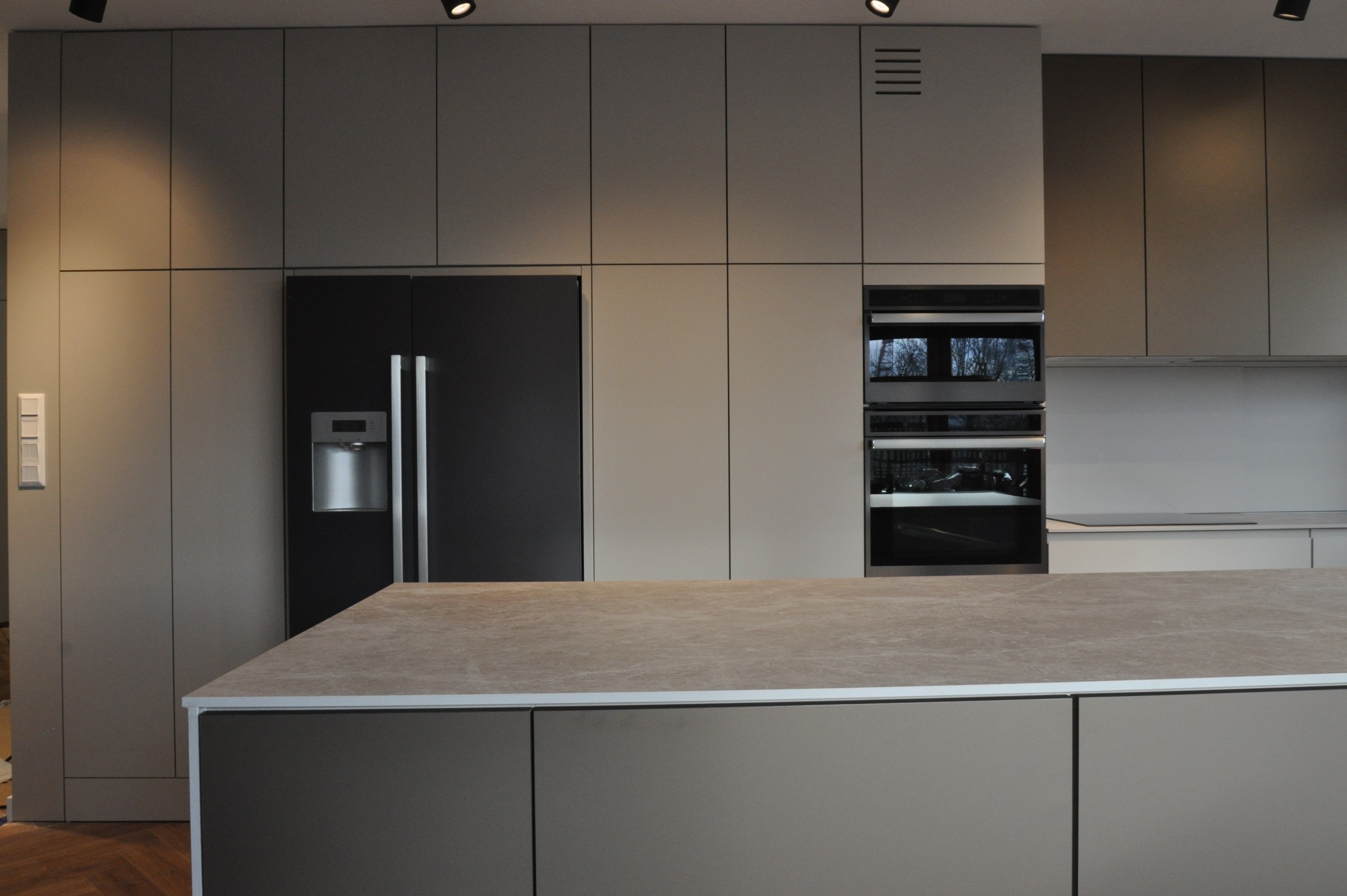 Nowoczesna kuchnia z szarymi frontami, czarną lodówką side-by-side i wyspą z blatem w kolorze piaskowym. Oświetlenie punktowe w suficie.