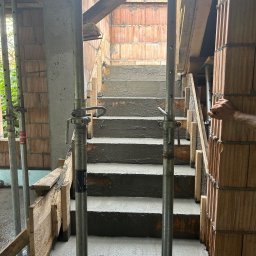 NIEBIESKIE KASKI SPÓŁKA Z OGRANICZONĄ ODPOWIEDZIALNOŚCIĄ - Wewnętrzne schody żelbetowe w trakcie budowy, z widocznym szalunkiem i podporami stalowymi. Ceglane ściany w tle, fragment ręki po prawej stronie.