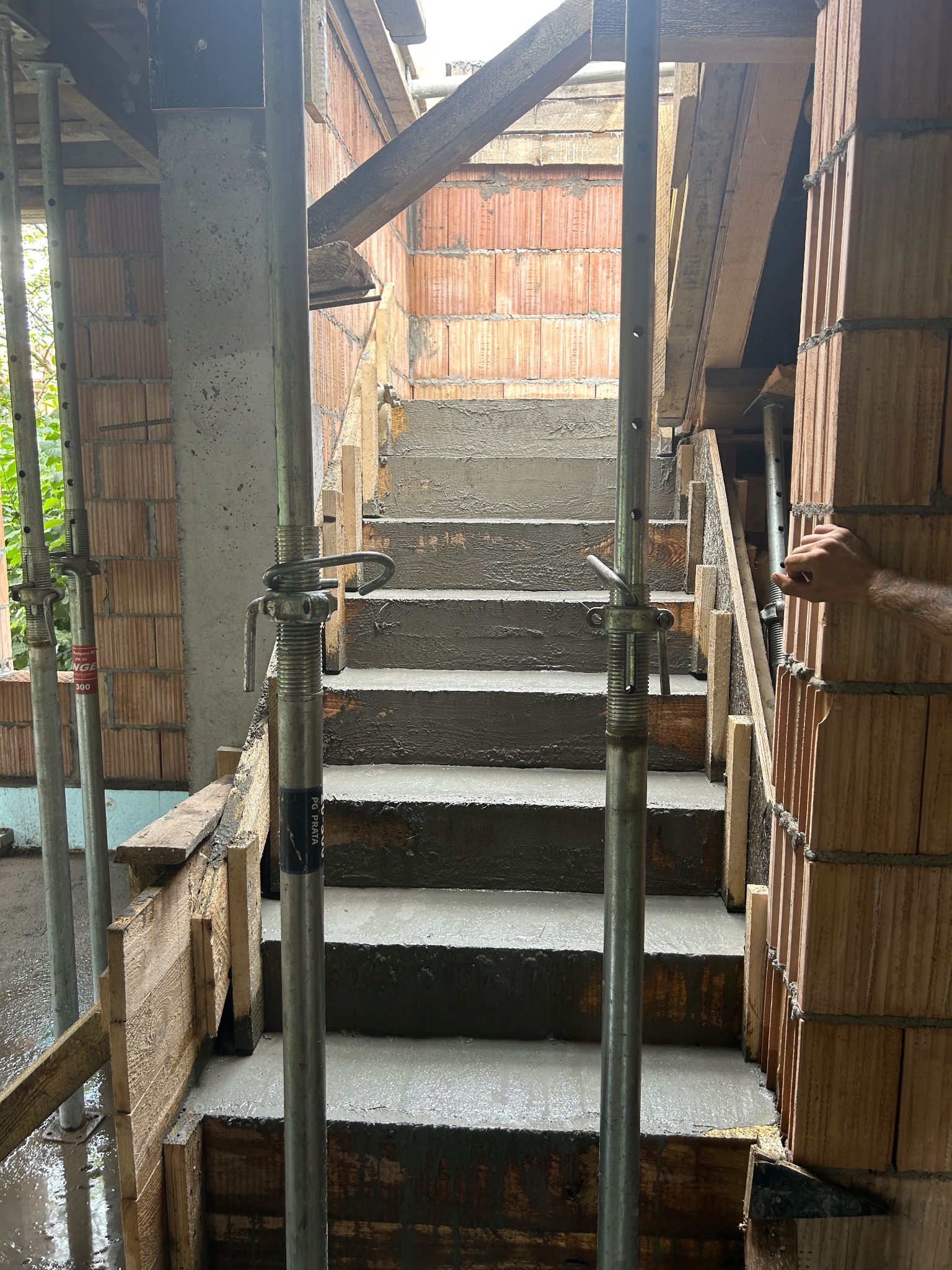 Wewnętrzne schody żelbetowe w trakcie budowy, z widocznym szalunkiem i podporami stalowymi. Ceglane ściany w tle, fragment ręki po prawej stronie.