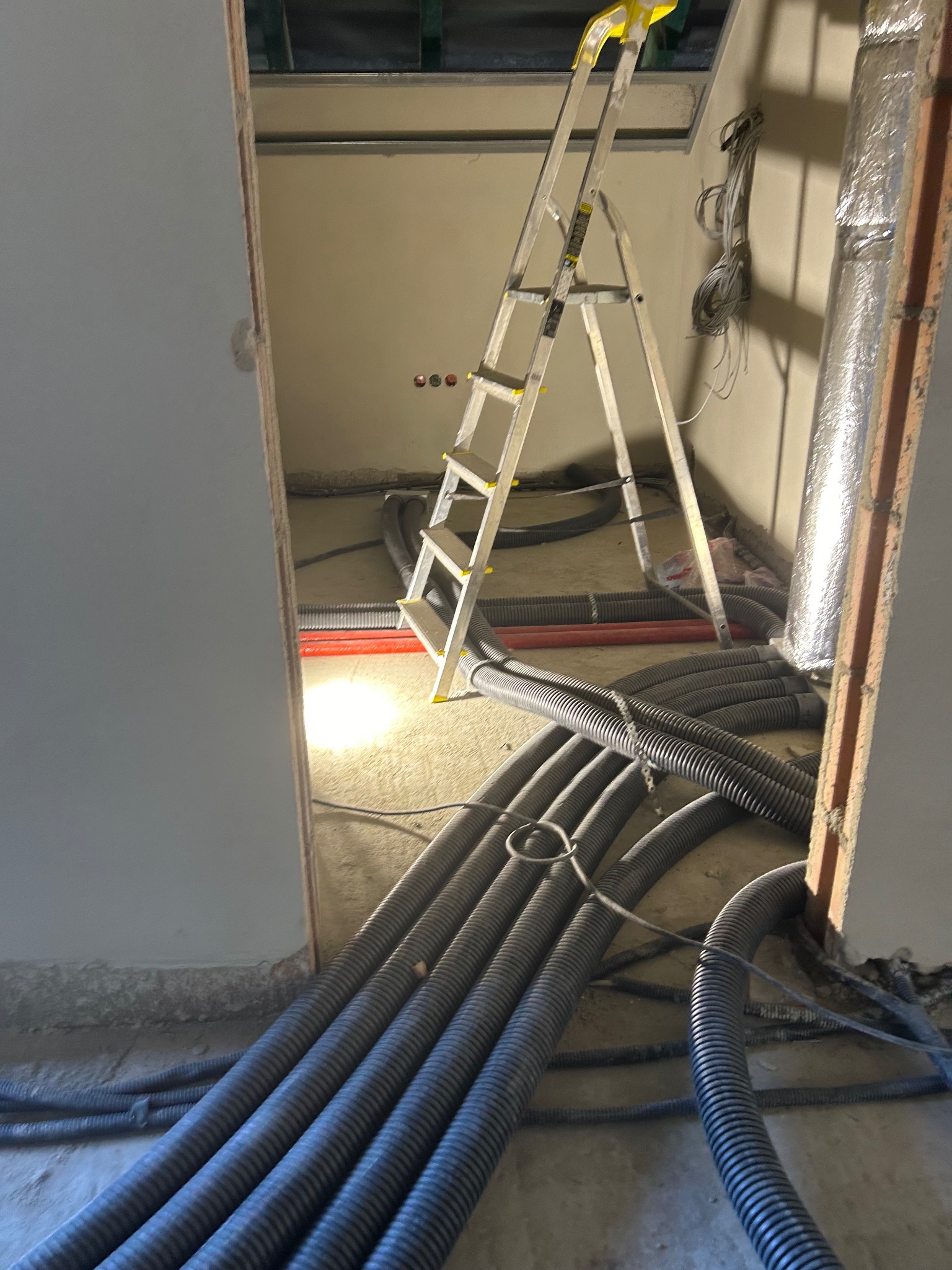 Instalacja rur karbowanych na podłodze w trakcie budowy, widoczna drabina i przewody. Ściany w surowym stanie, widok zza krawędzi ściany.