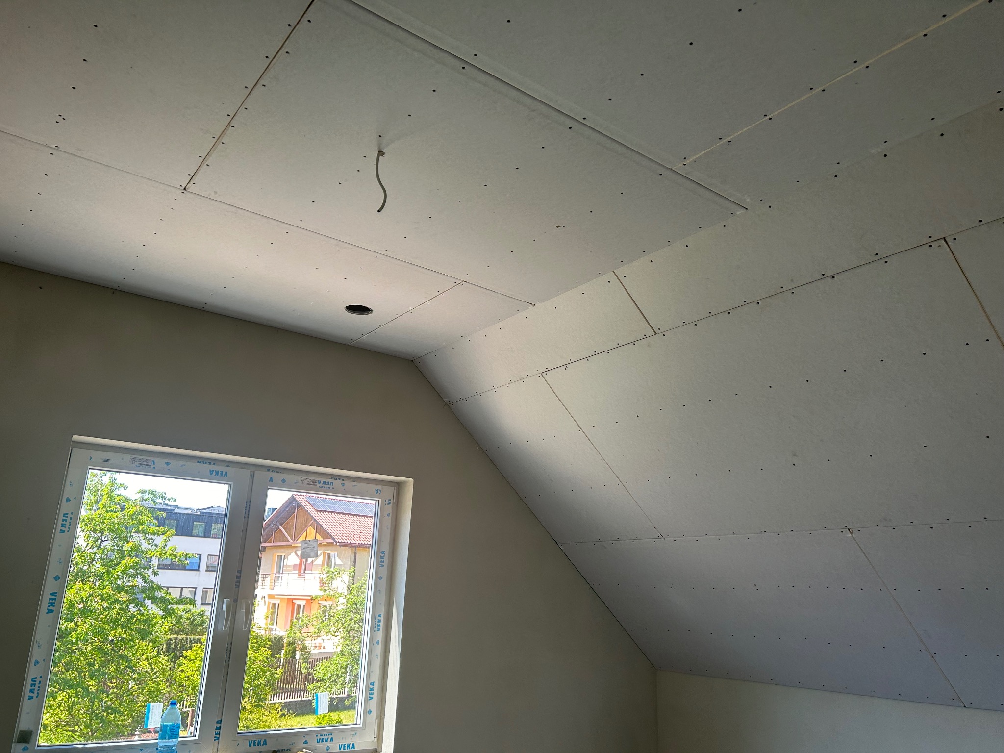 Poddasze z oknem po montażu płyt gipsowo-kartonowych na skośnym suficie. Widoczne łączenia płyt i otwór na oświetlenie. Na parapecie butelka wody.