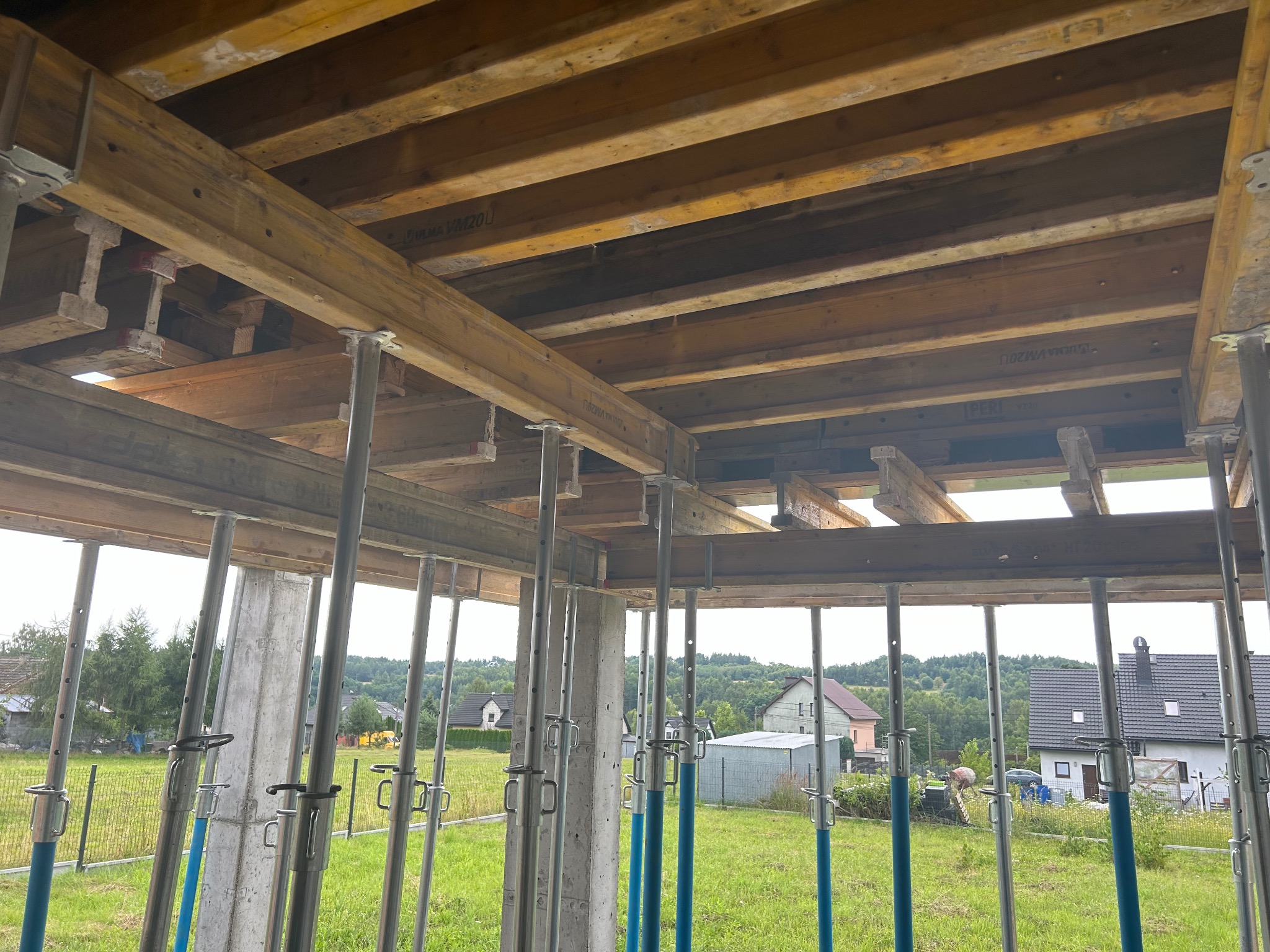 Szlifowanie betonu: szalunek stropu na podporach teleskopowych, betonowy słup nośny. Widok na zielone otoczenie i zabudowę jednorodzinną.