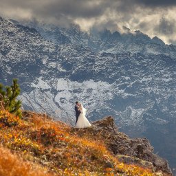 Julita Chudko Photography - Para młoda w objęciach na tle górskim w jesiennej scenerii. Suknia panny młodej kontrastuje z surowym krajobrazem. Romantyczne ujęcie z pleneru.