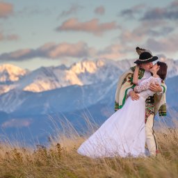 Julita Chudko Photography - Para młoda w strojach góralskich całuje się na tle gór, oświetlonych zachodzącym słońcem. Widok na Tatry, trawa, ciepłe barwy.
