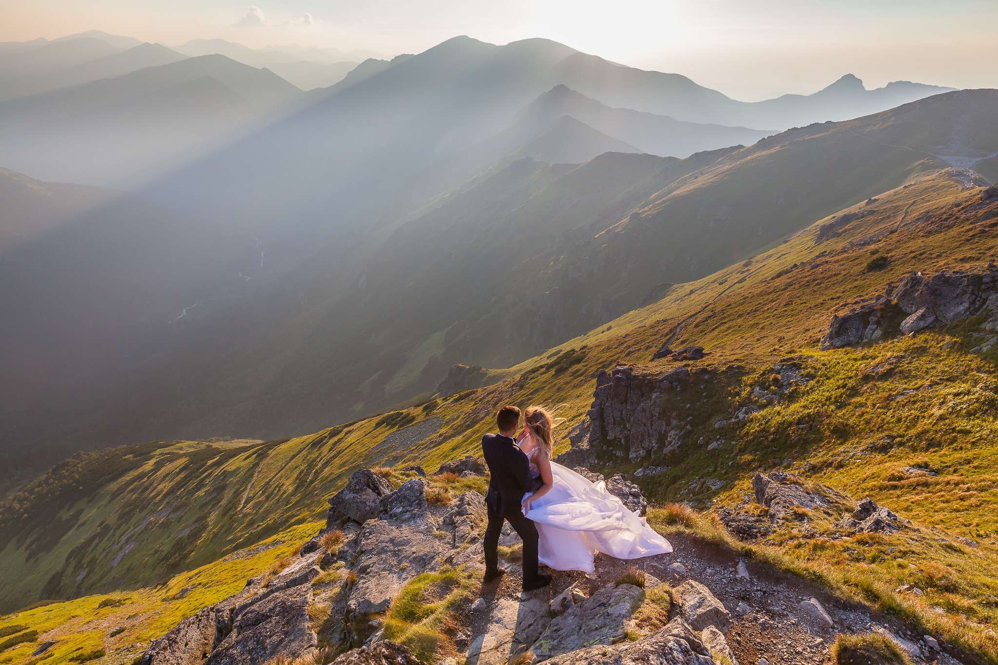 Para młoda na tle gór oświetlonych promieniami słońca. Suknia powiewa na wietrze, tworząc malowniczy efekt. Widok z góry na pasmo górskie w oddali.