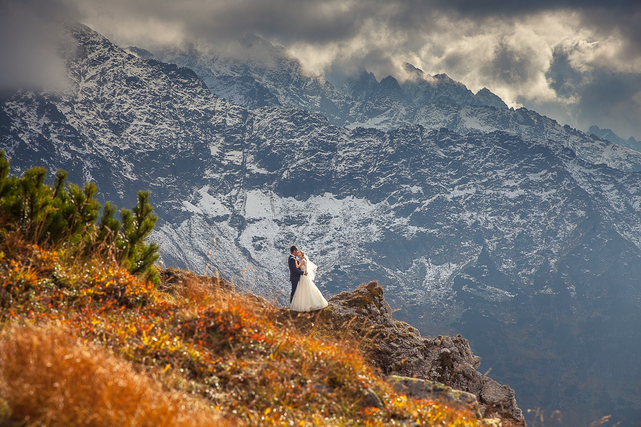 Para młoda w objęciach na tle górskim w jesiennej scenerii. Suknia panny młodej kontrastuje z surowym krajobrazem. Romantyczne ujęcie z pleneru.