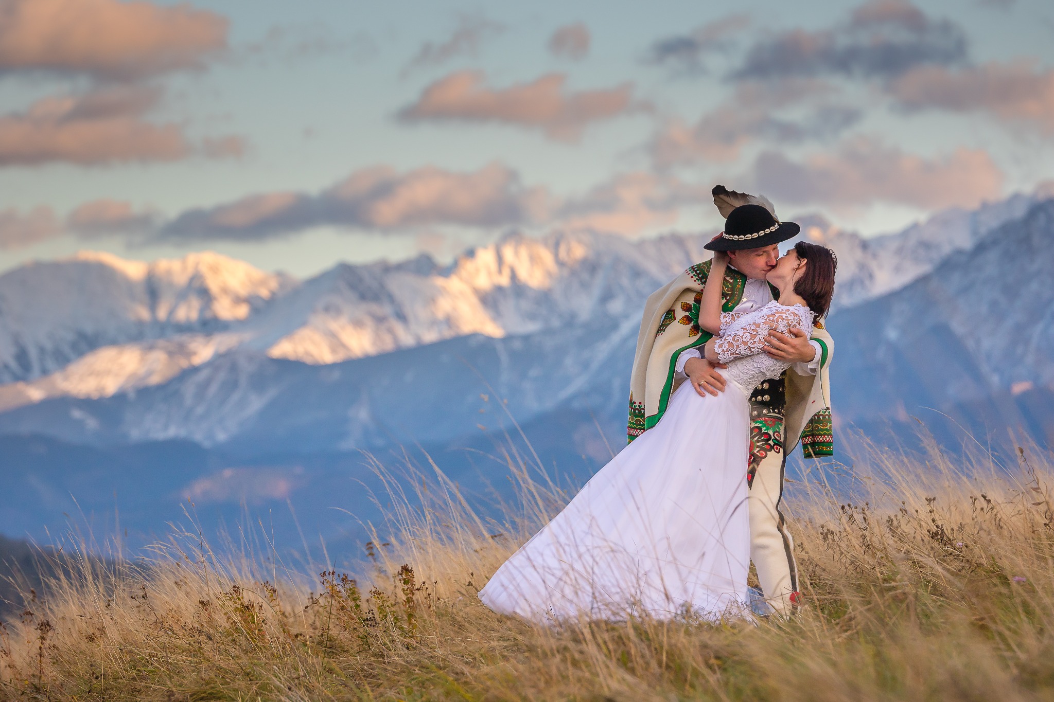 Para młoda w strojach góralskich całuje się na tle gór, oświetlonych zachodzącym słońcem. Widok na Tatry, trawa, ciepłe barwy.