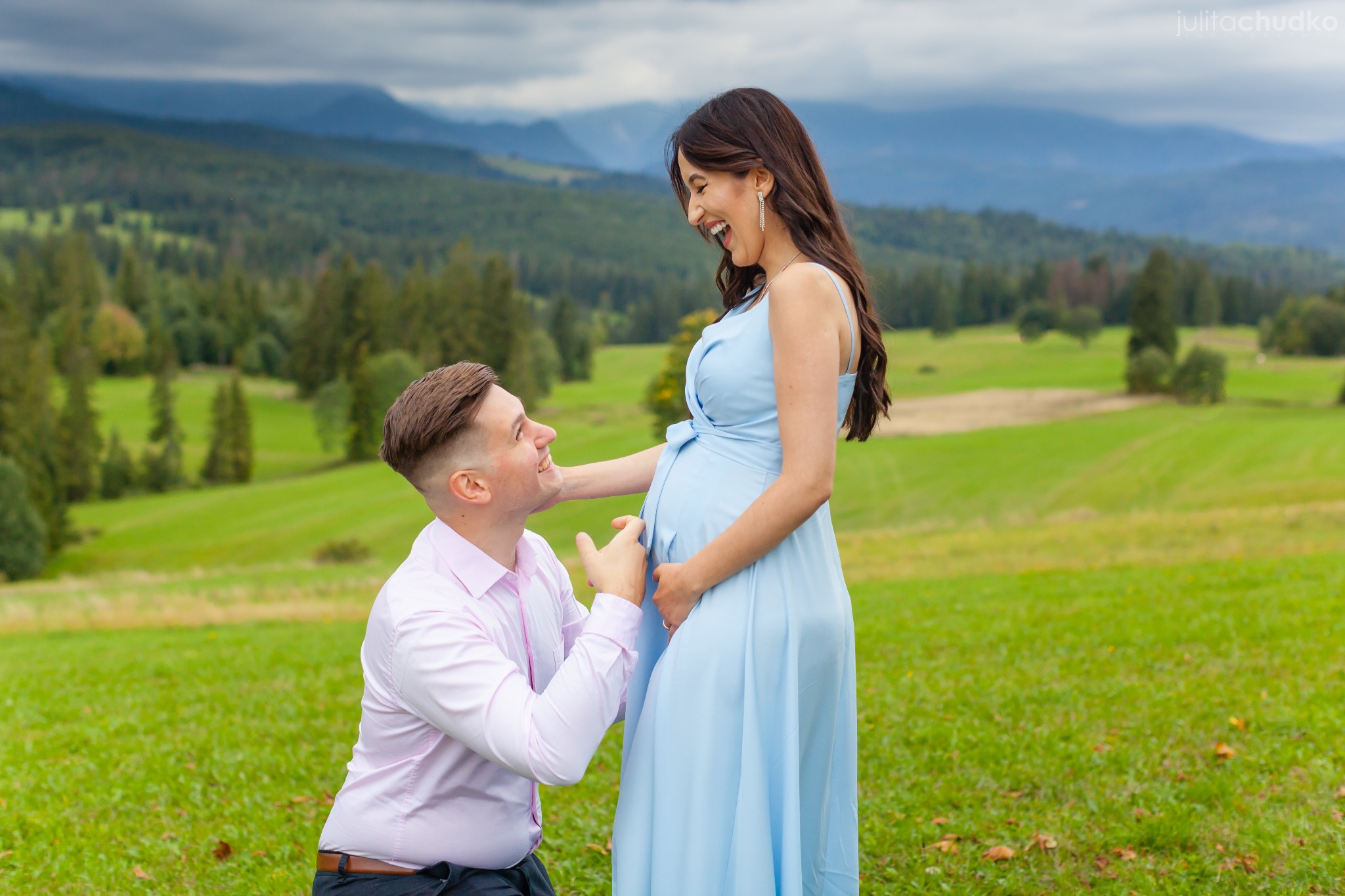 Uśmiechnięta kobieta w błękitnej sukni ciążowej i klęczący mężczyzna na tle górskiego krajobrazu. Sesja w plenerze, naturalne światło.