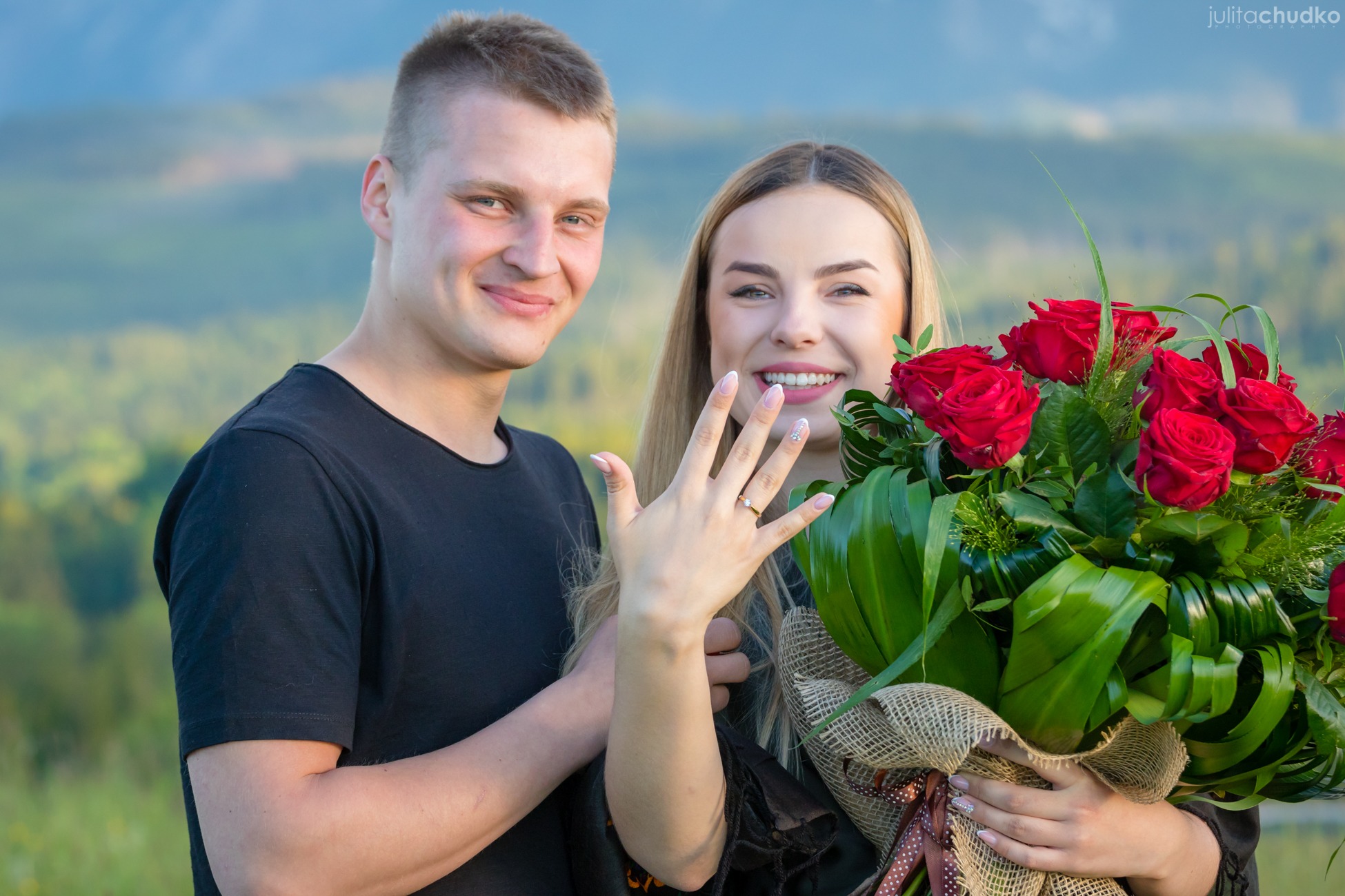 Uśmiechnięta para z bukietem czerwonych róż na tle gór. Kobieta prezentuje pierścionek zaręczynowy, dłoń z pierścionkiem na pierwszym planie.