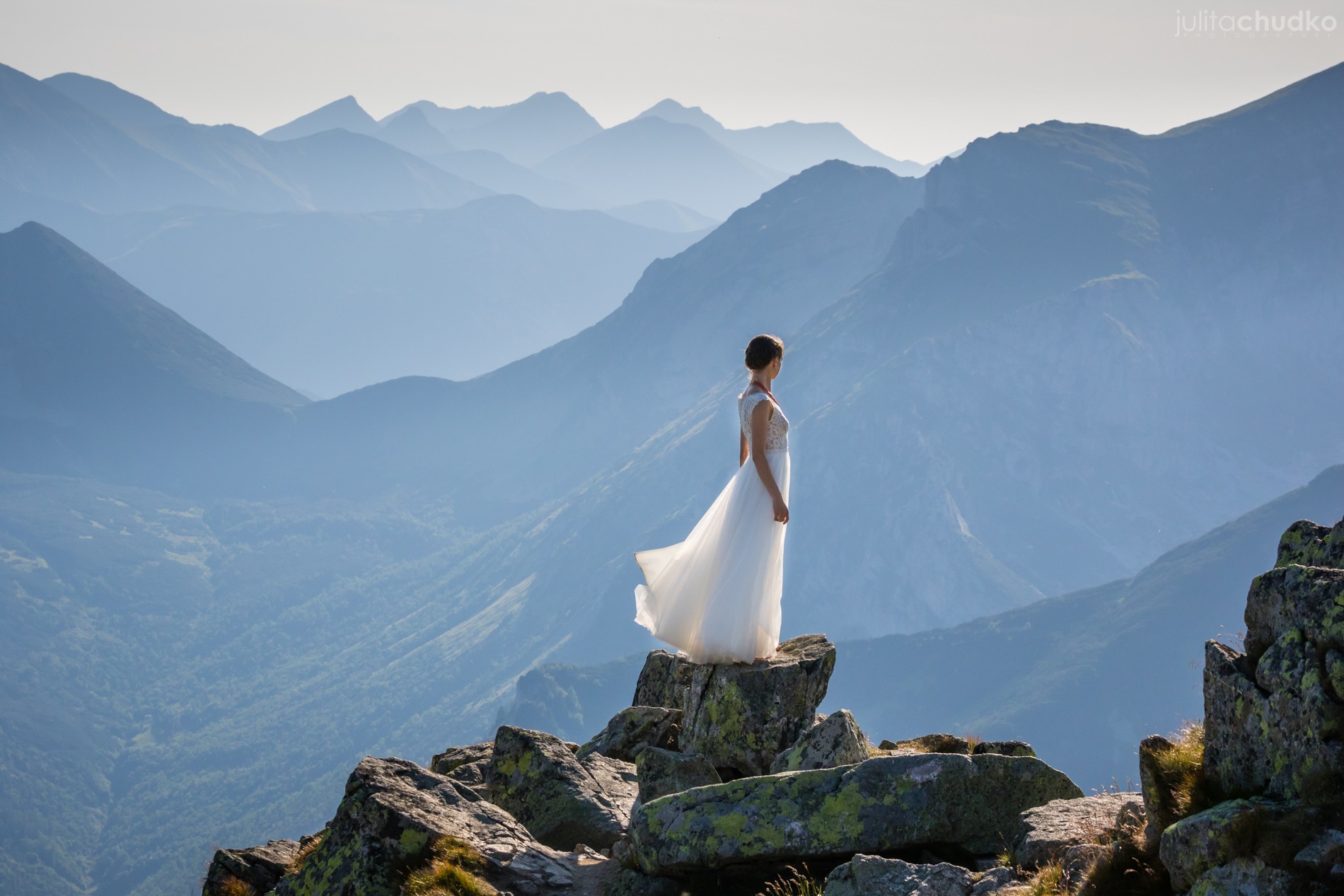 Panna młoda w zwiewnej sukni stoi na szczycie skalistego urwiska z widokiem na góry spowite mgłą. Ujęcie z dołu podkreśla majestat krajobrazu i delikatność sukni.