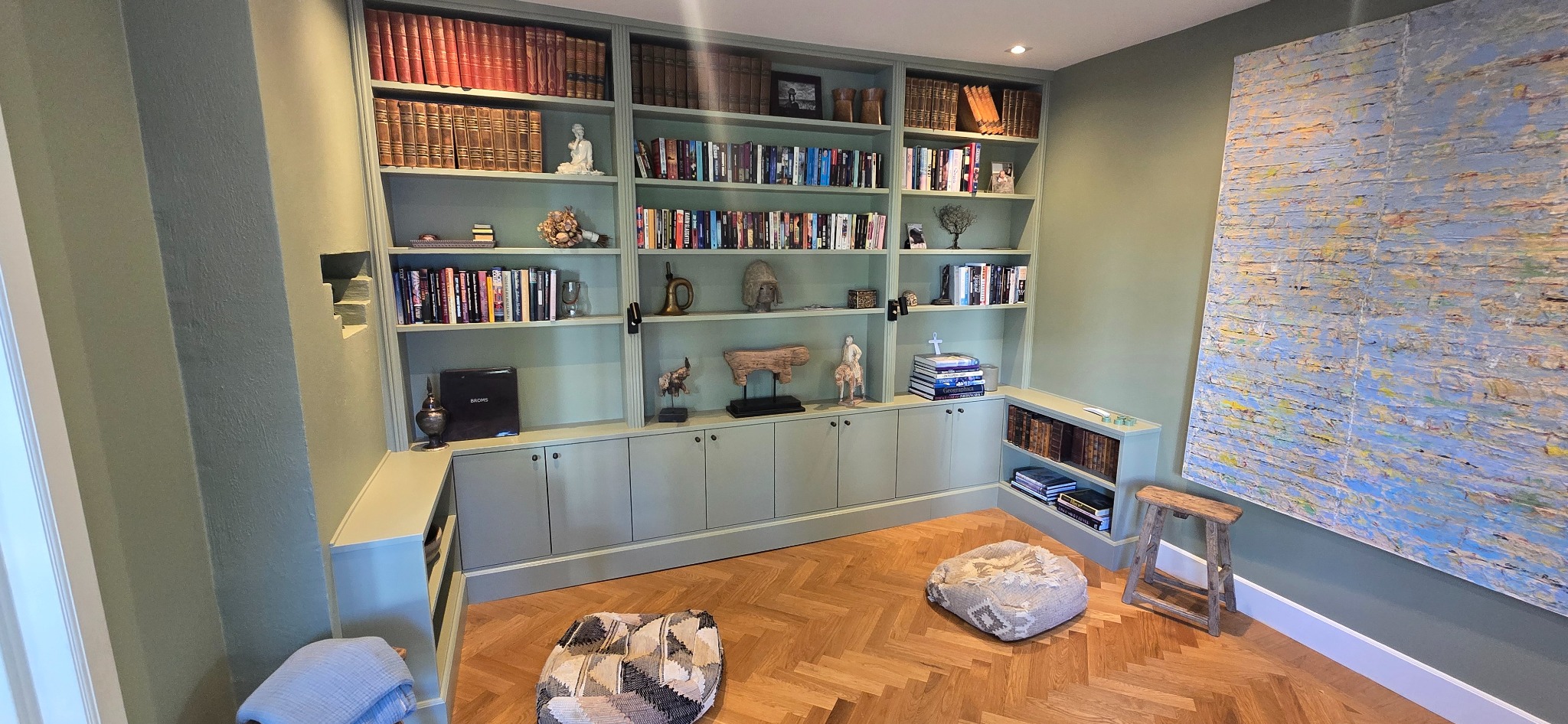 Zabudowa biblioteczna w odcieniach zieleni, z półkami pełnymi książek i dekoracji. Parkiet w jodełkę, pufy i obraz dopełniają wnętrze.