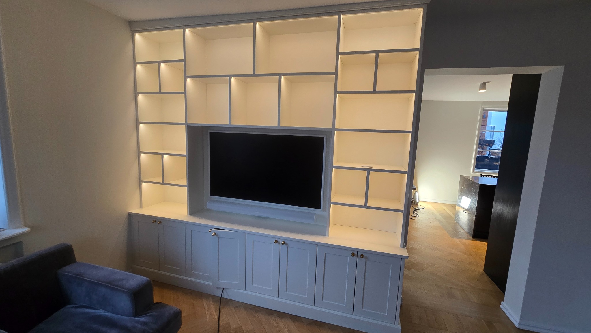 Biała zabudowa meblowa z podświetlanymi półkami i miejscem na telewizor, widok z perspektywy salonu, parkiet w jodełkę na podłodze, widok na inne pomieszczenie.