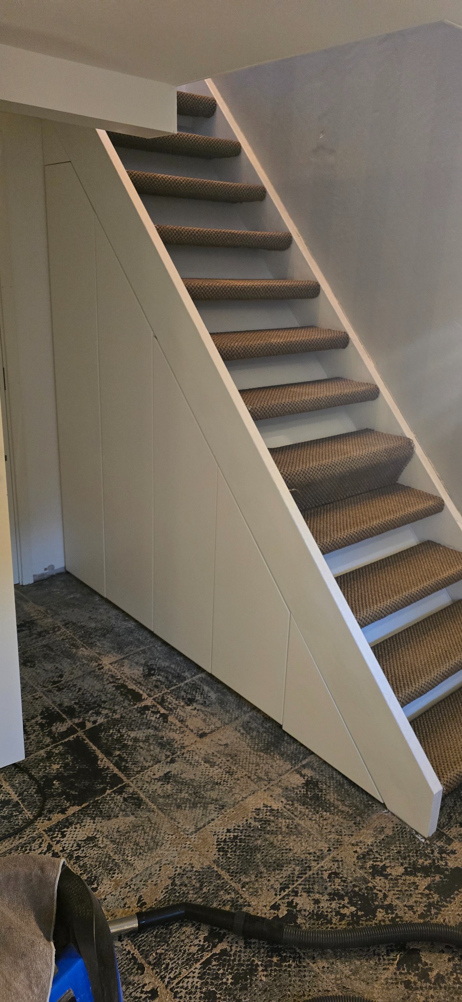 Białe schody z wykładziną dywanową i zabudową pod schodami. Widoczny fragment podłogi z wzorem i odkurzacz. Ujęcie z dołu, podkreślające konstrukcję.