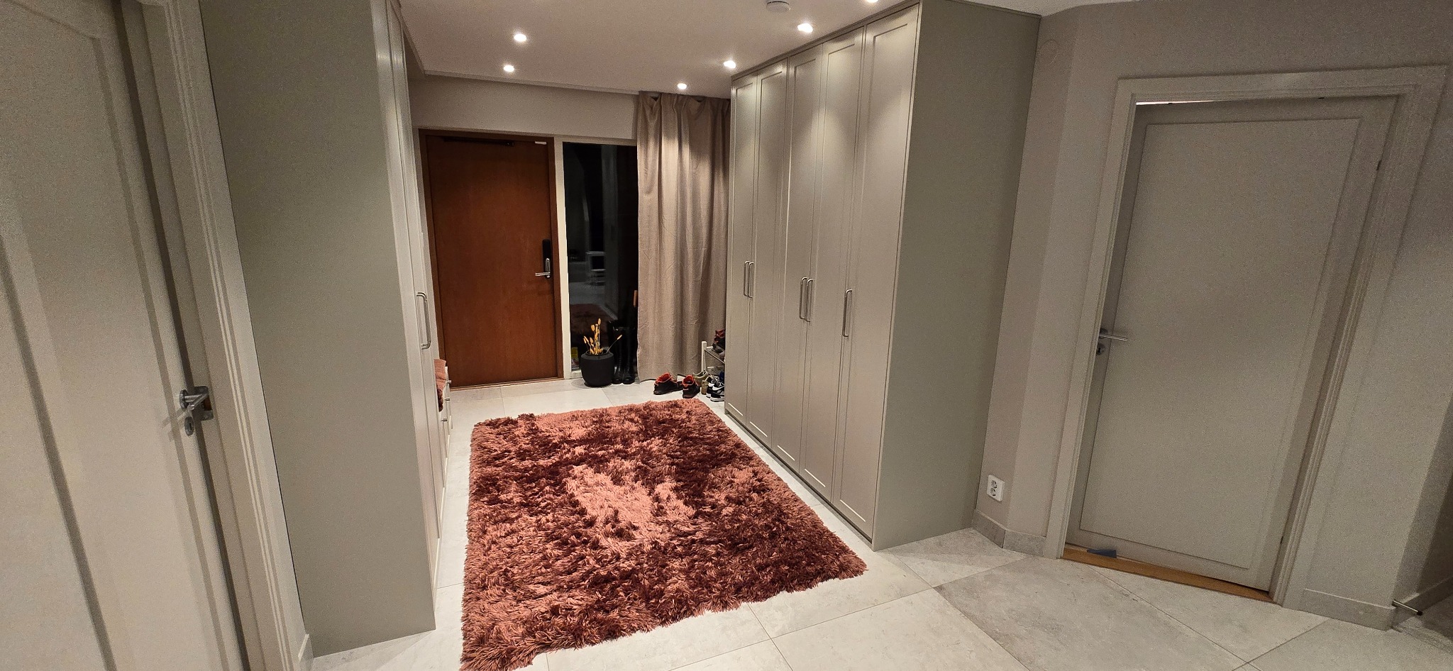 Jasne wnętrze przedpokoju z beżową szafą w zabudowie, brązowym dywanem i dwiema parami drzwi. Widoczne oświetlenie sufitowe i dekoracje przy drzwiach wejściowych.
