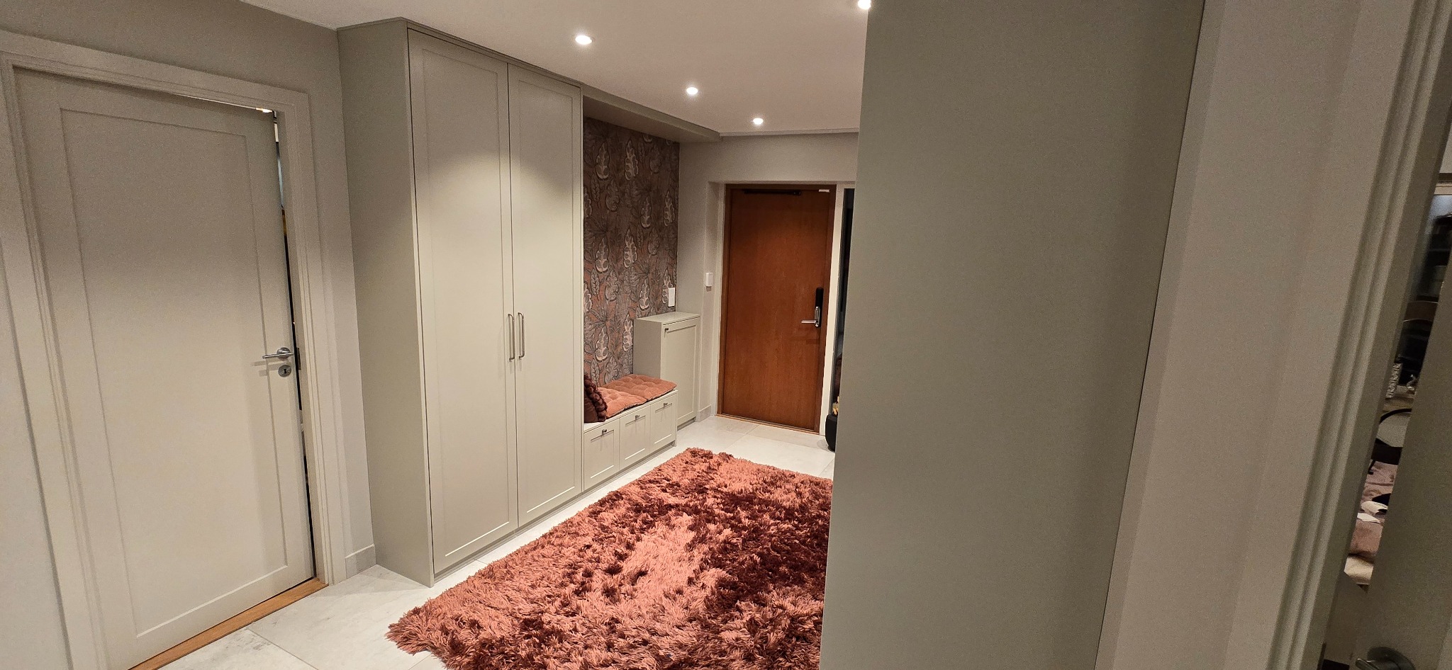 Jasne wnętrze przedpokoju z szafą w zabudowie, siedziskiem i wzorzystą tapetą. Na podłodze puszysty, różowy dywan. Widoczne drzwi wejściowe i wewnętrzne.