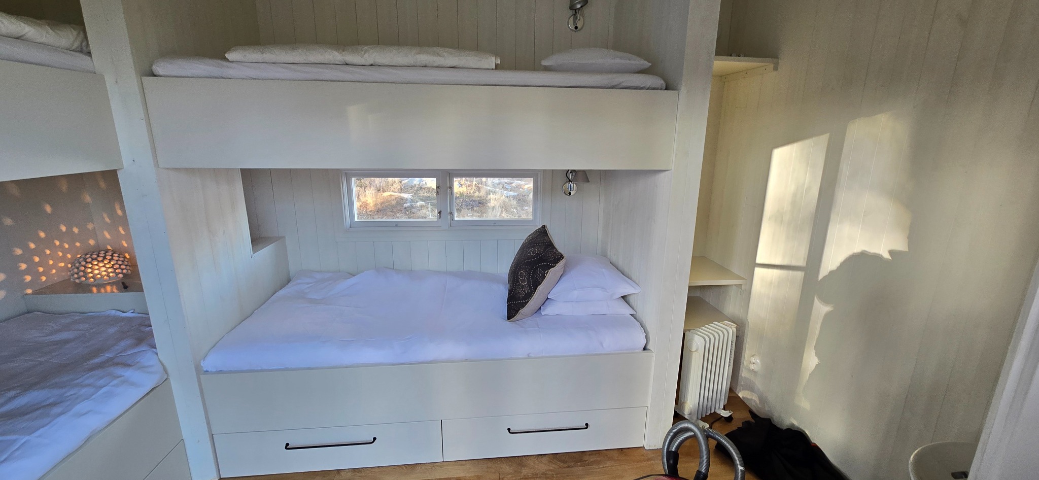 Białe łóżko piętrowe z szufladami w pokoju wykończonym boazerią. Widoczne okno z widokiem na zewnątrz i lampka nocna na półce. Minimalistyczny design.