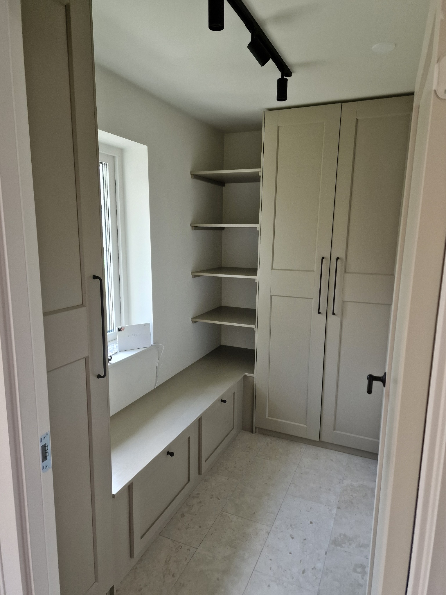 Jasne wnętrze z zabudową meblową: szafa z długimi uchwytami, półki wnękowe, siedzisko pod oknem z szufladami. Stonowana kolorystyka, punktowe oświetlenie sufitowe.