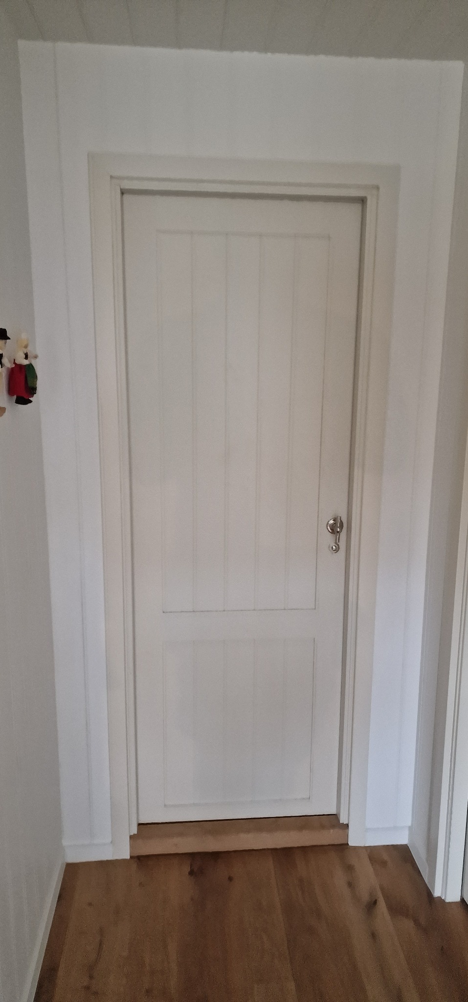 Białe, dwuczęściowe drzwi wewnętrzne z srebrną klamką, osadzone w białej futrynie, na tle drewnianej podłogi. Widoczne detale wykończenia ścian i listew przypodłogowych.