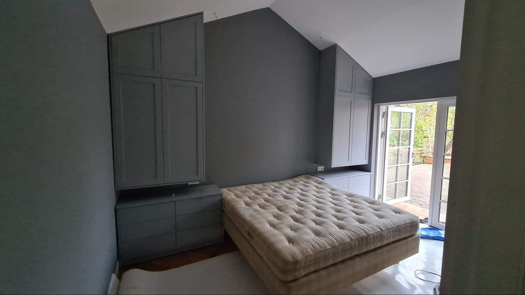 Szara sypialnia z zabudową meblową pod skosami w Krobi. Szafy z szufladami i szafkami wiszącymi. Materac na podłodze, drzwi balkonowe z widokiem na zieleń.