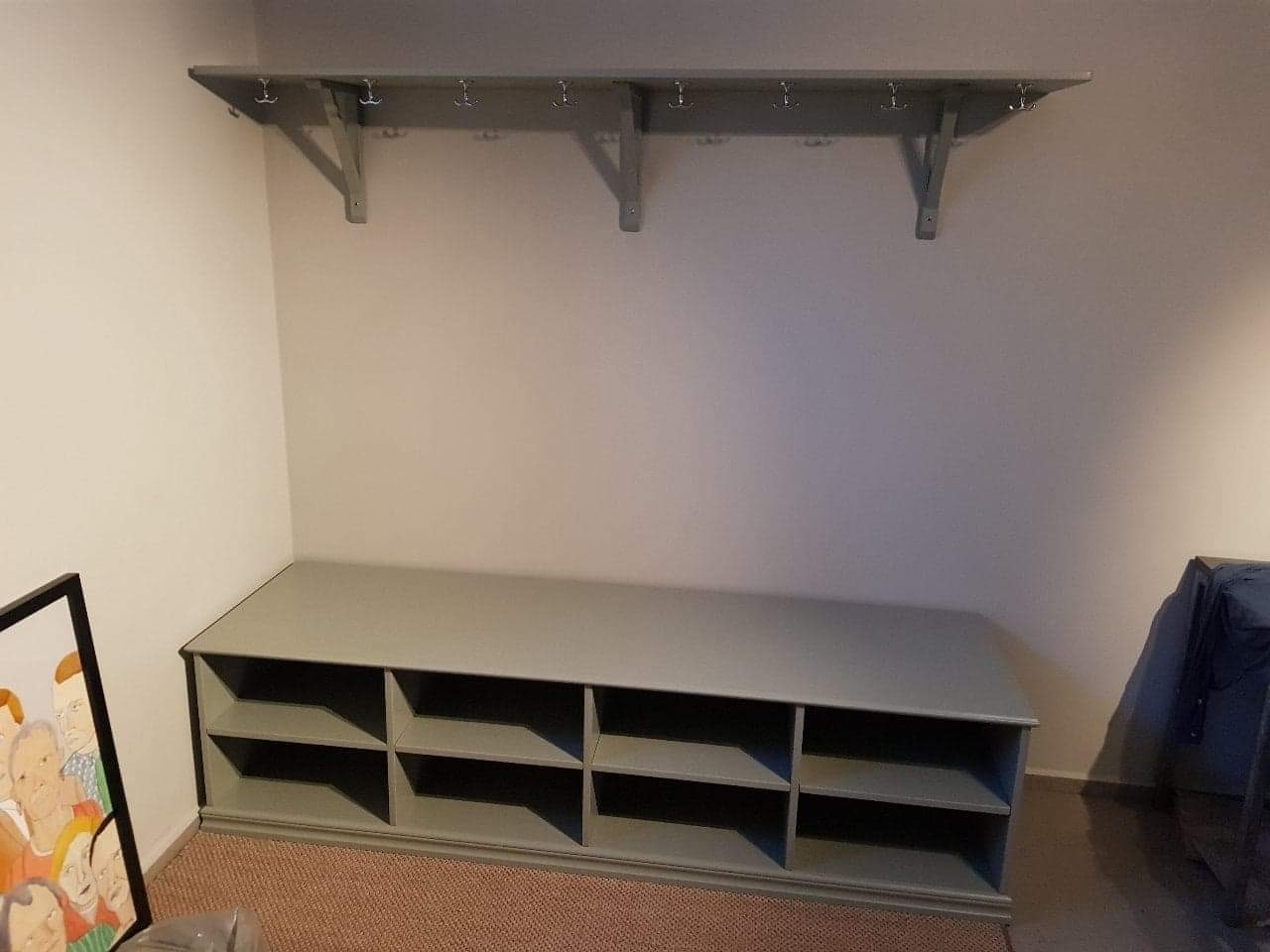 Szara szafka na buty z wieszakiem w przedpokoju. Widoczne półki na obuwie i rząd haczyków na ubrania. Prosty, minimalistyczny design, jasne ściany.