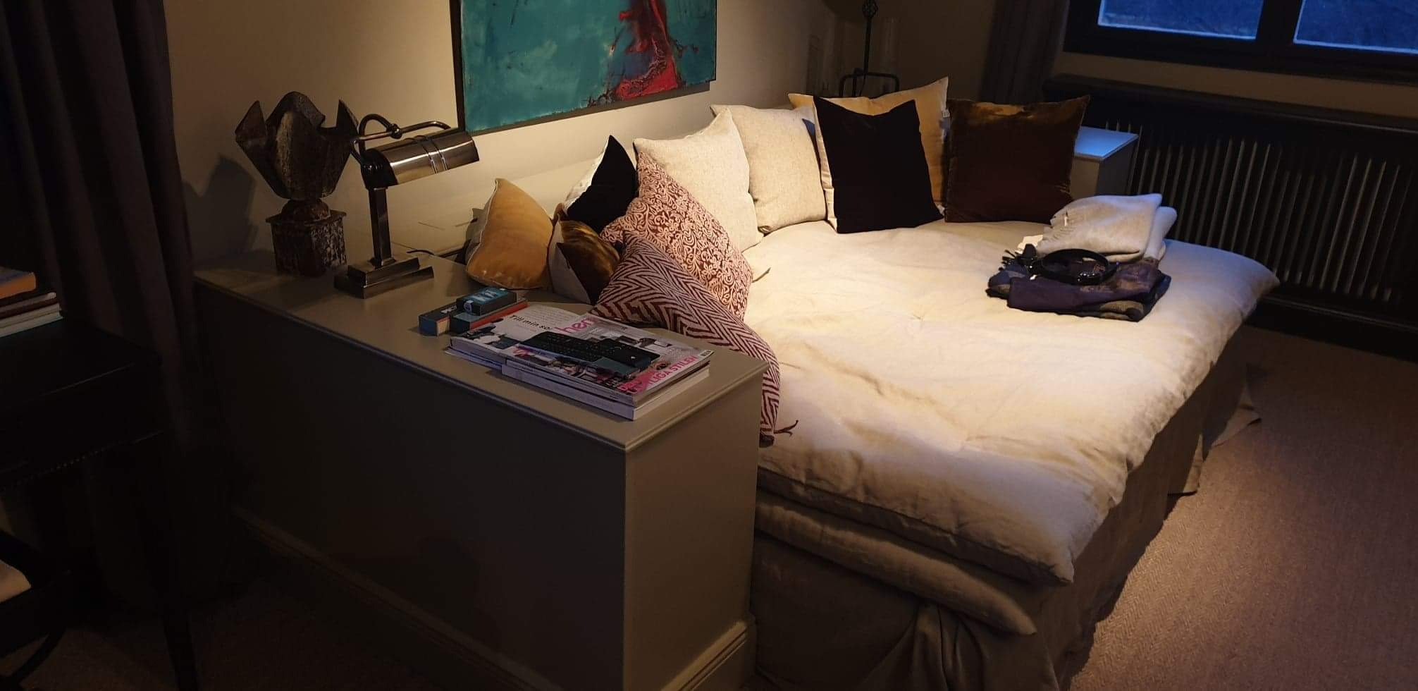 Aranżacja sypialni z łóżkiem, stolikiem nocnym z lampką i dekoracjami. Na stoliku leżą czasopisma i pilot. Nad łóżkiem wisi obraz.