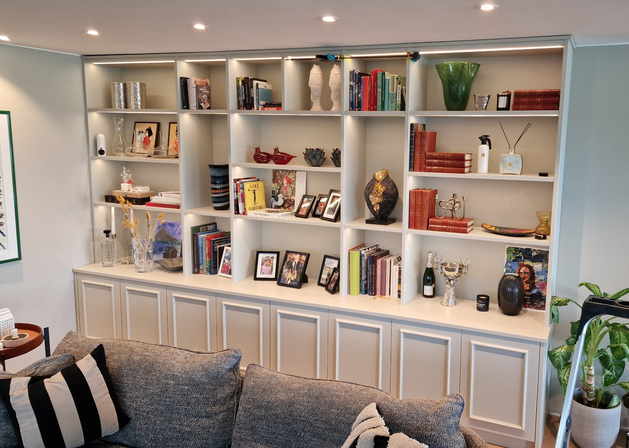 Biały regał z oświetleniem LED, wypełniony książkami i dekoracjami, widoczny zza szarej sofy z poduszkami w salonie. Element wykończenia wnętrza.