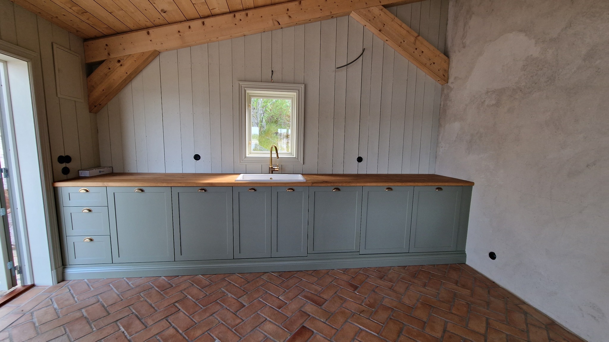 Kuchnia w stylu rustykalnym z drewnianym sufitem i ceglaną podłogą. Szare szafki z drewnianym blatem i złotą armaturą. Widoczne elementy instalacji elektrycznej.