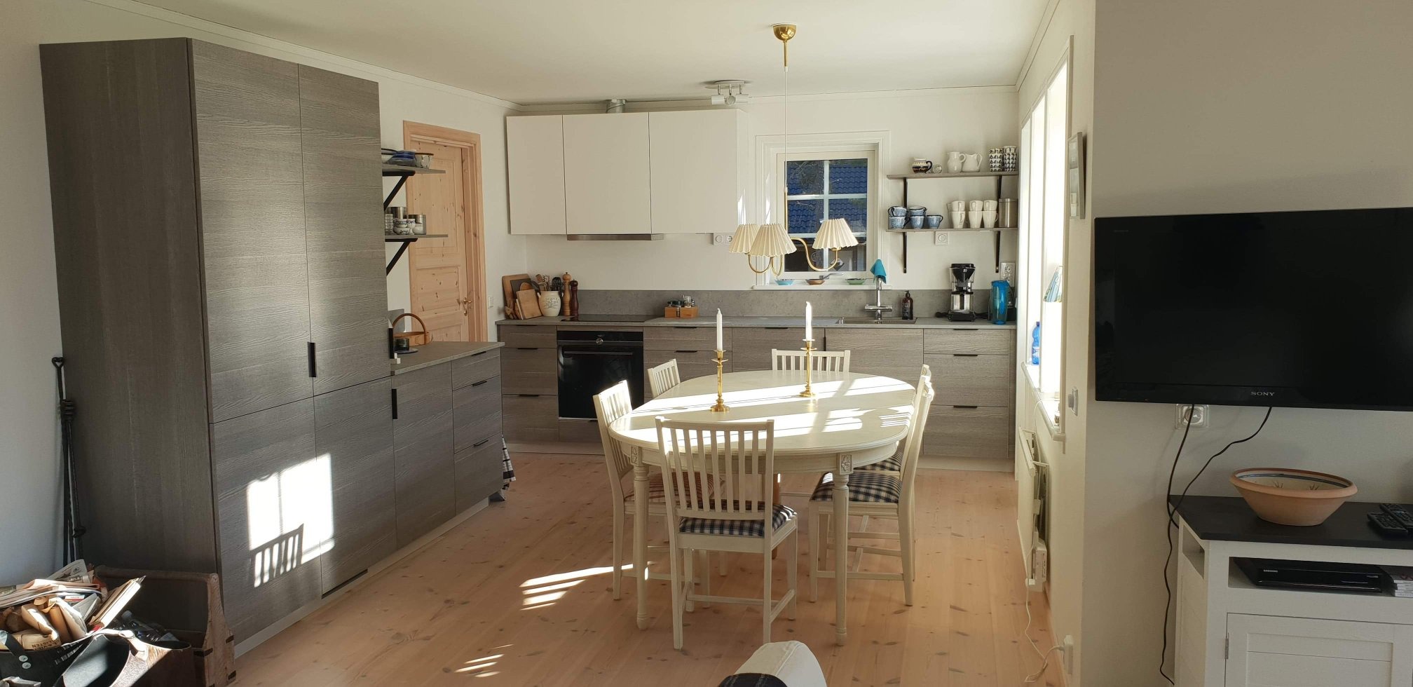 Jasna kuchnia z drewnianą podłogą, szarymi szafkami i białymi górnymi szafkami. Okrągły stół z krzesłami w centrum, oświetlony naturalnym światłem.