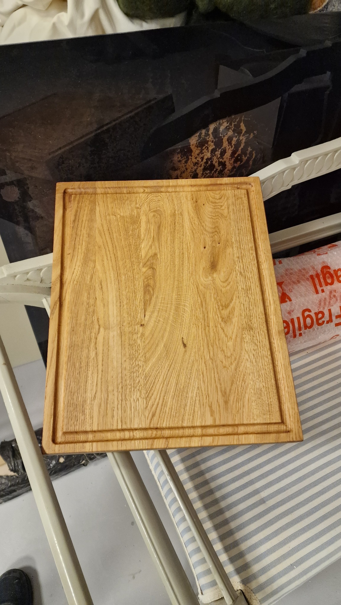 Jasna, kwadratowa deska z drewna dębowego z frezowanym obramowaniem, leżąca na krześle z niebiesko-białym obiciem. W tle widoczny blat z ciemnego kamienia.