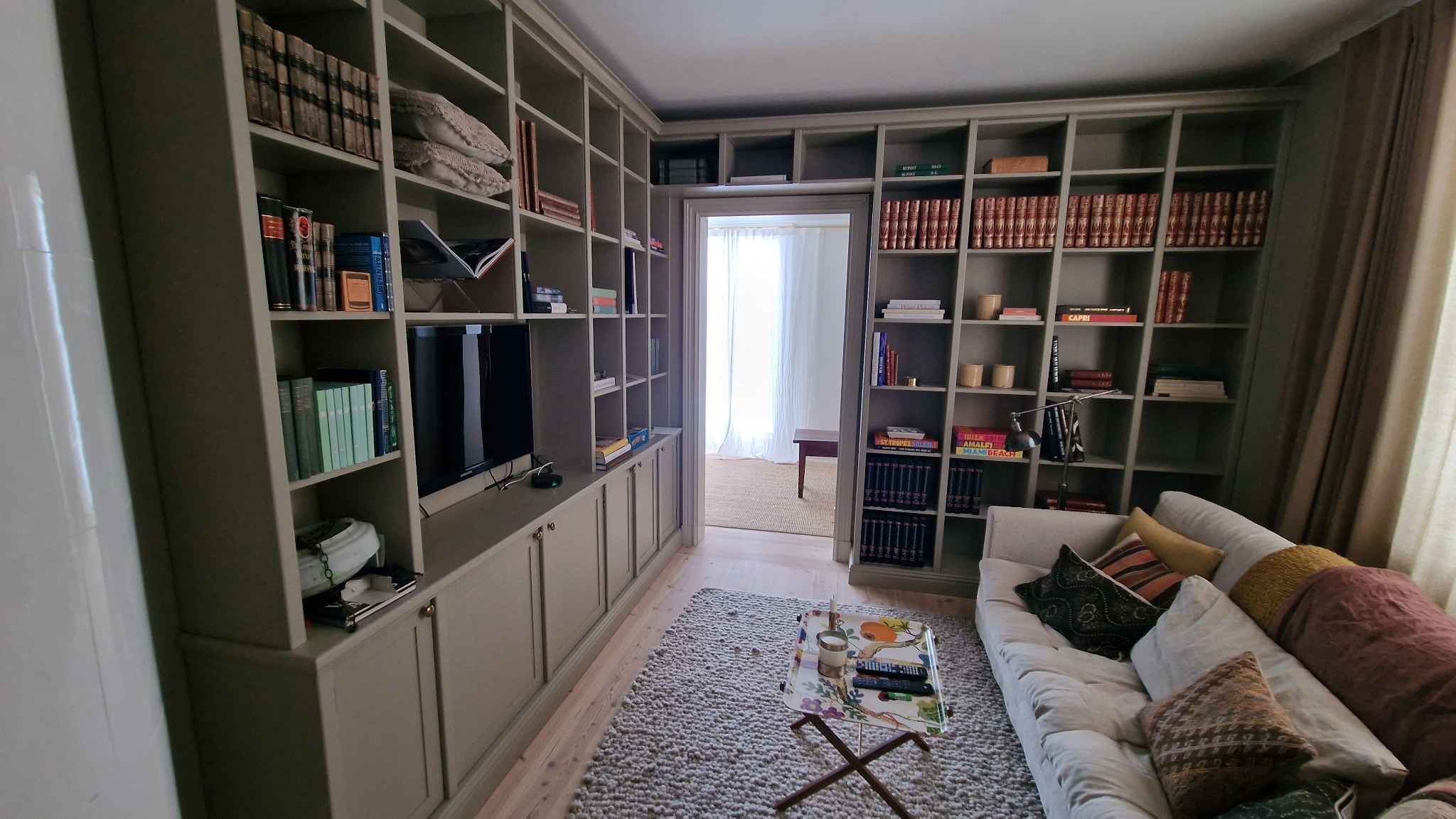 Zabudowa biblioteczna w salonie z szarymi regałami na książki i dekoracje, z telewizorem wkomponowanym w mebel. Dywan w stylu skandynawskim i sofa z poduszkami.