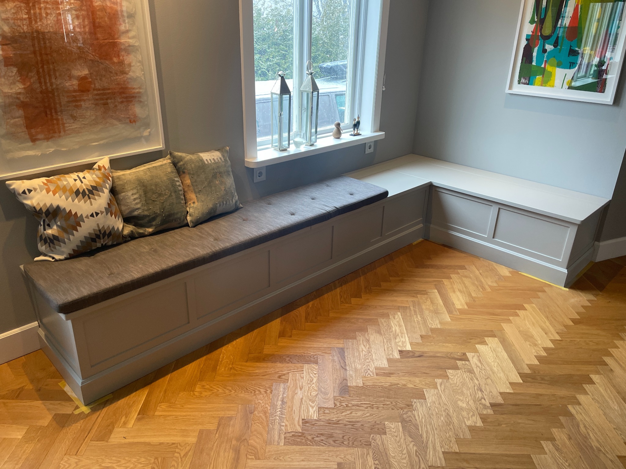 Wnęka okienna z siedziskiem i poduszkami w szarościach, parkiet w jodełkę. Dekoracje okienne i obrazy na ścianach dopełniają aranżację wnętrza.