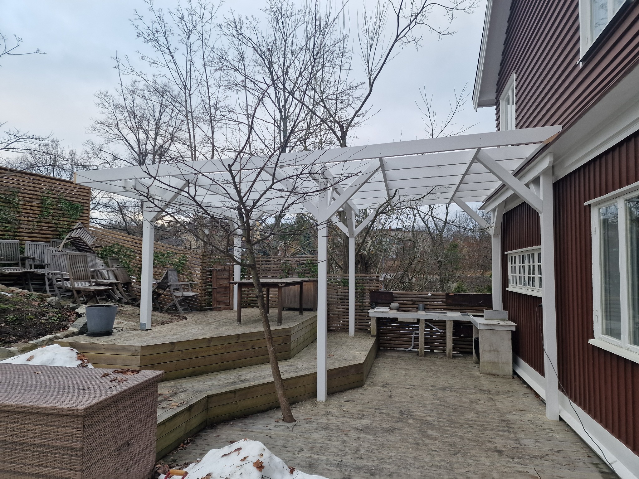 Drewniany taras z białą pergolą przy domu o brązowej elewacji. Widoczne meble ogrodowe i drzewa bez liści. Zimowy krajobraz z resztkami śniegu.
