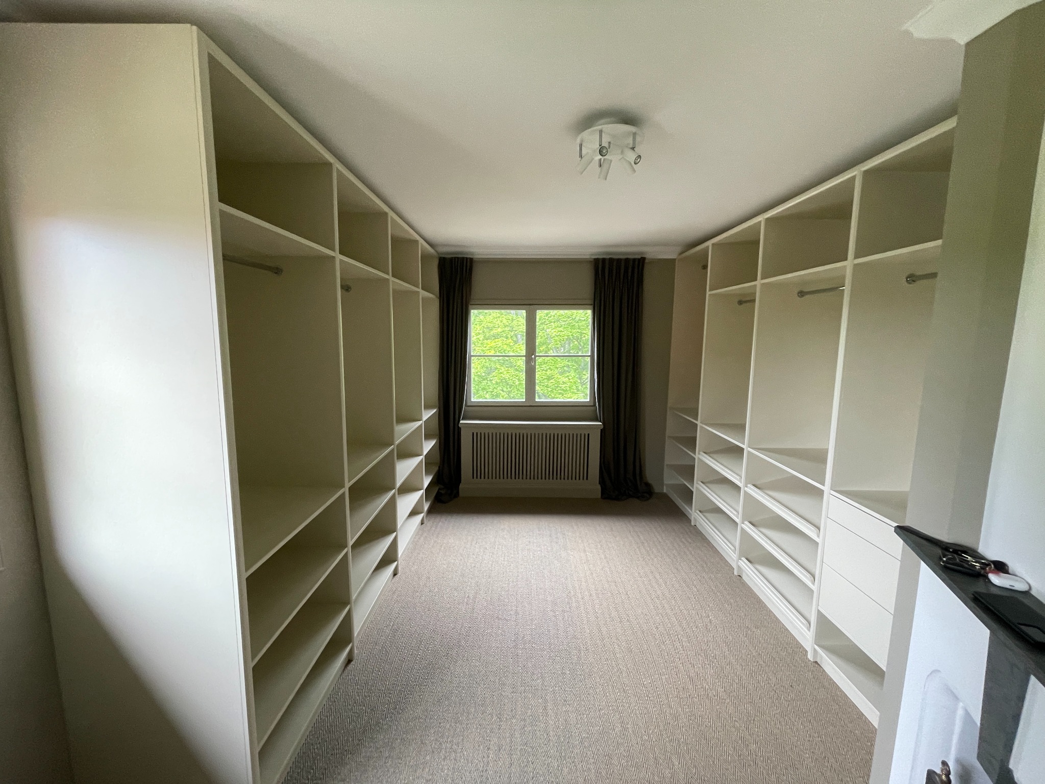 Biała, zabudowana garderoba z półkami i drążkami, widok na okno z zasłonami. Jasna podłoga i sufit. Minimalistyczny design, symetryczny układ szaf.