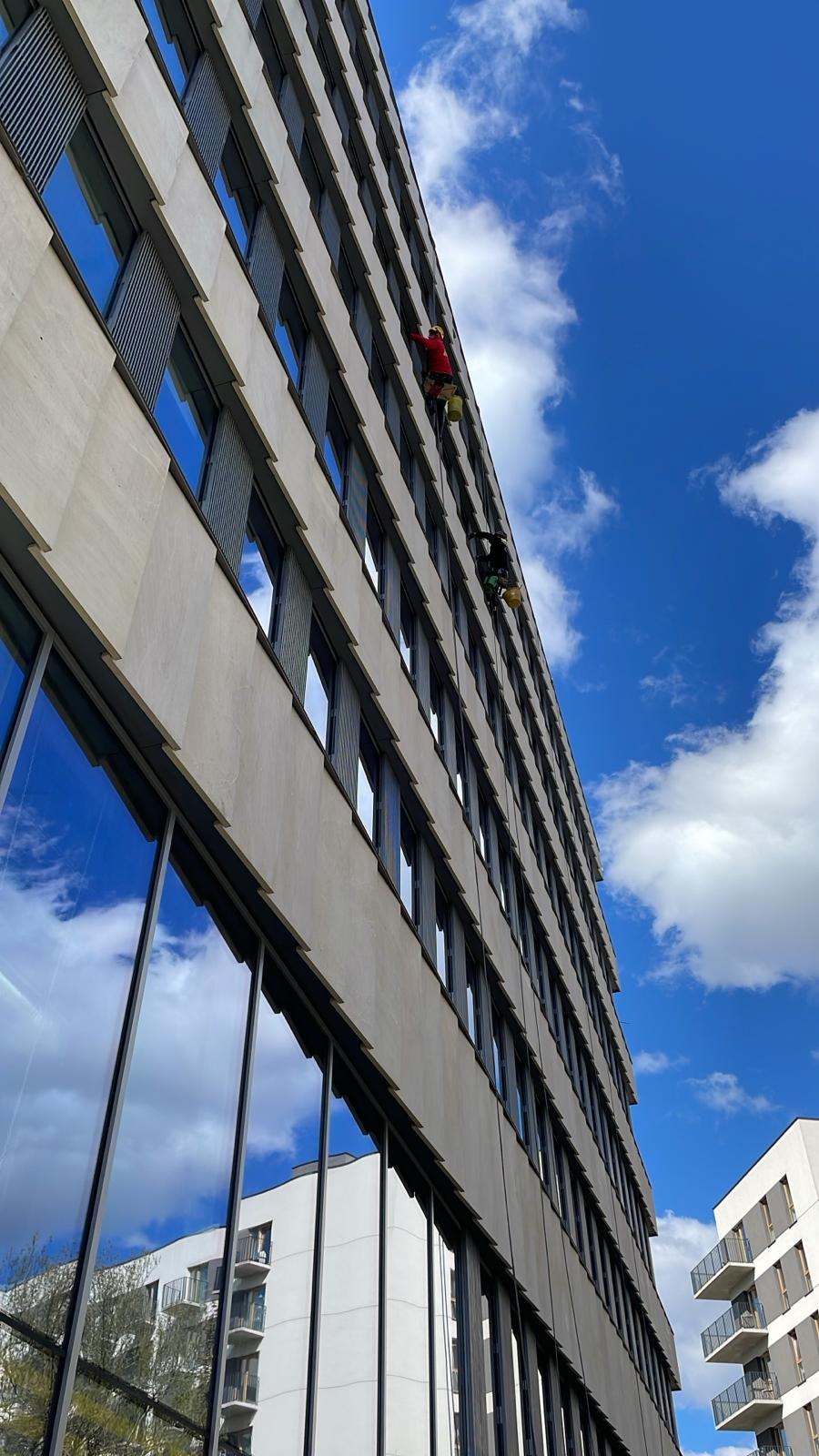 Dwóch alpinistów w uprzężach, myjących okna na wysokościowcu. Błękitne niebo z chmurami odbija się w szybach. Widok z dołu, budynek biurowy.