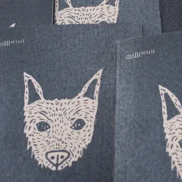 Stos wizytówek/ulotek z szarą okładką, na której widnieje grafika głowy psa w stylu linorytu i logo 'millprint'. Minimalistyczny design, stonowana kolorystyka.