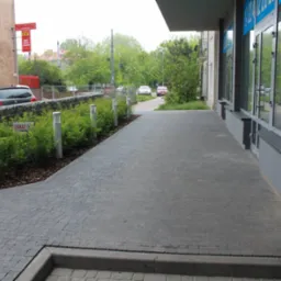 Szara kostka brukowa ułożona wzdłuż budynku z oknami, oddzielona od zieleni ozdobnej niskimi lampami. Widoczny fragment ulicy i zaparkowane samochody w tle.