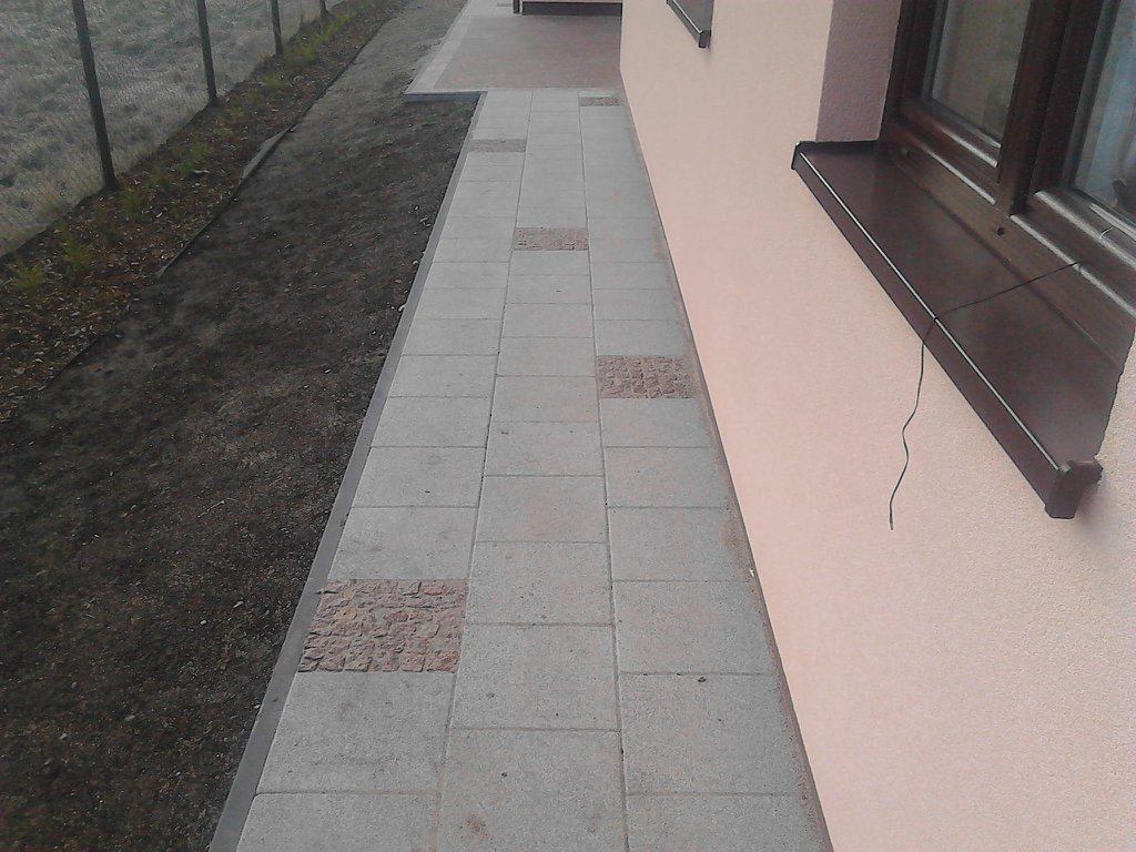 Ścieżka z szarej kostki brukowej z dekoracyjnymi wstawkami z kostki granitowej, biegnąca wzdłuż ściany budynku i rabaty ziemnej.