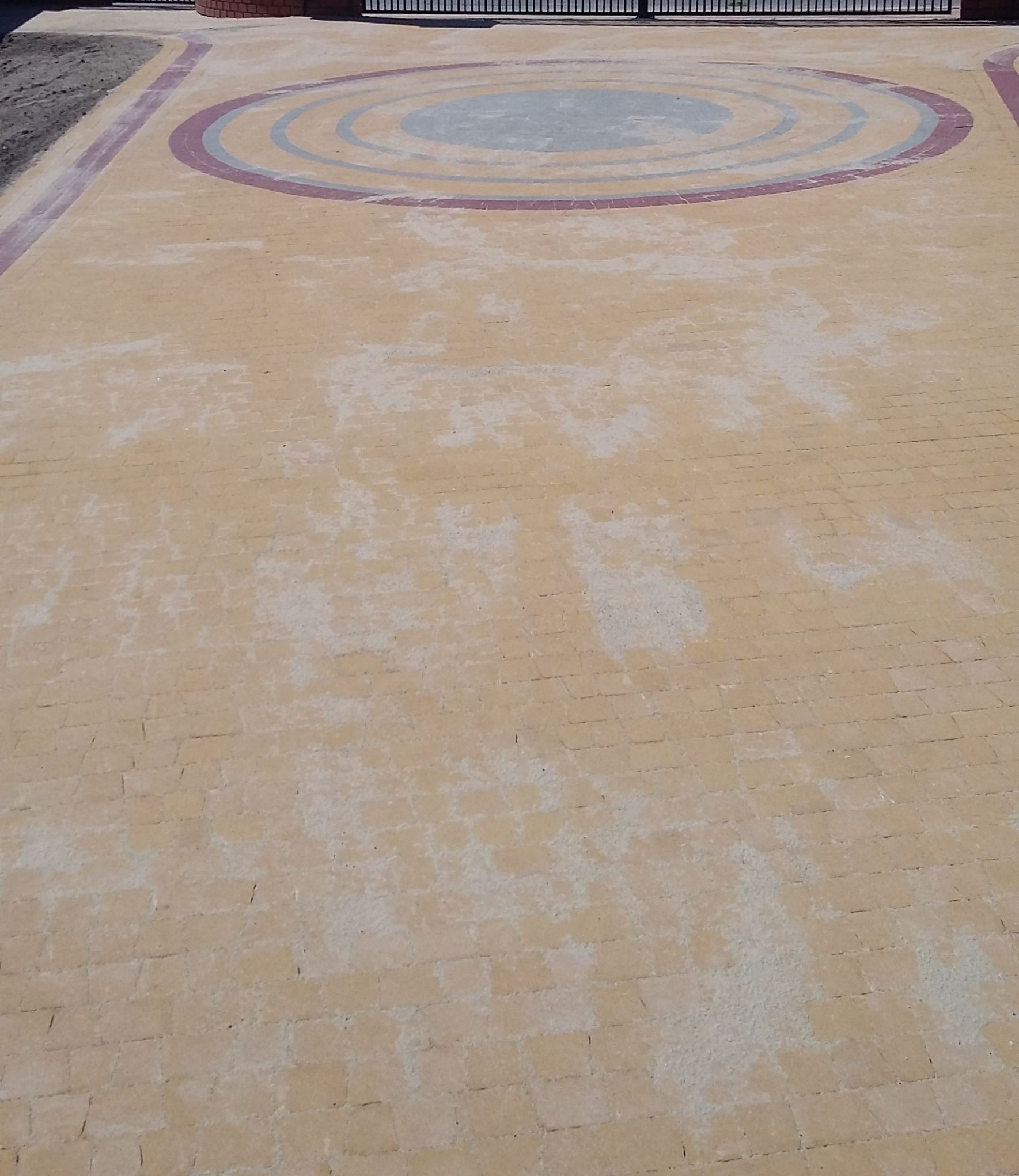 Beżowa kostka brukowa z dekoracyjnym wzorem koncentrycznych okręgów w odcieniach szarości i czerwieni, widziana z góry, prowadząca do czarnej bramy.