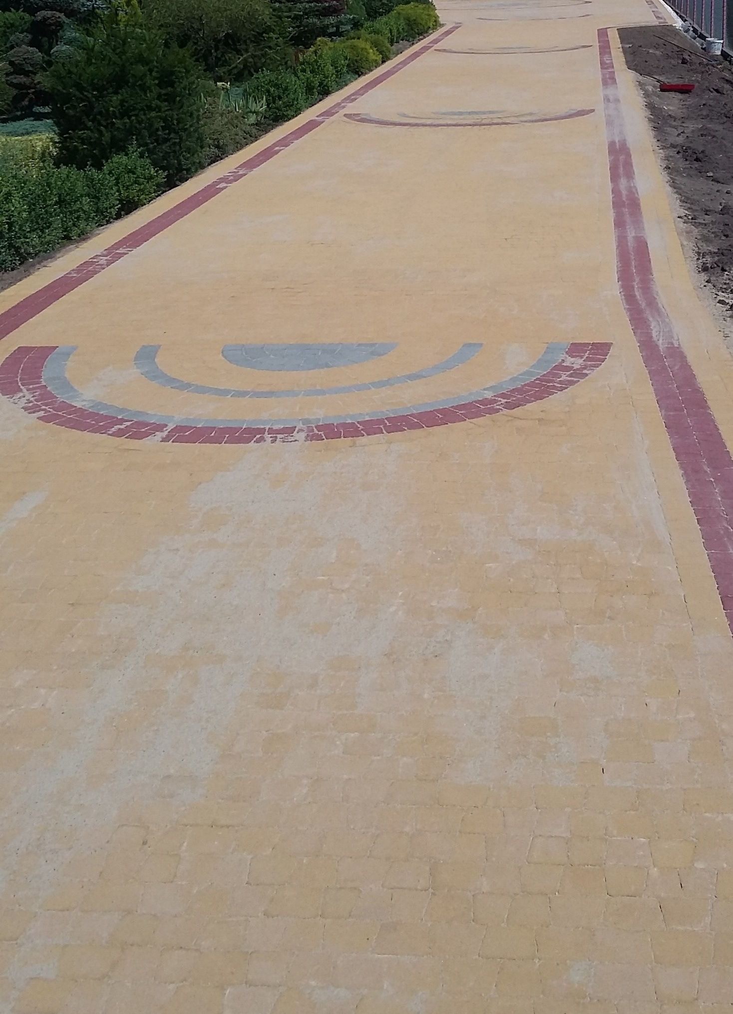 Długa, wybrukowana ścieżka z wzorami w kształcie półksiężyców z czerwonej i szarej kostki, otoczona zielenią i świeżo nasypaną ziemią.